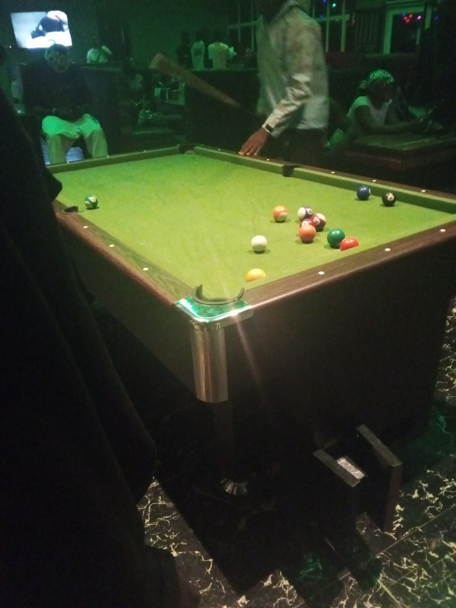 The image captures an indoor scene, likely a bar or recreation hall in Lokoja, Nigeria, during the evening or night, indicated by the dim, artificial green lighting. The central focus is a green-felted pool table, with several pool balls scattered across its surface, including a white cue ball, orange, red, green, blue, black, and striped balls.
Several people are visible. A person in a light-colored, long-sleeved shirt stands near the pool table, with their right hand resting on its edge, seemingly engaged in a game of pool. Another person, wearing light-colored pants and a dark top, is seated in the left background, while a person with a head covering is visible further right. Other blurry figures populate the dimly lit background. A pool cue is partially visible, suggesting ongoing play.
On the wall in the background, a television screen displays what appears to be a soccer match or sports event. The floor features dark, reflective tiles with a distinctive white marbling pattern. The corner pocket of the pool table has a metallic, possibly chrome, finish. A large dark fabric, likely part of a person's attire or an object close to the camera, occupies the left foreground, partially obscuring the view. The atmosphere seems social and relaxed, centered around the game of pool and possibly watching sports. No visible text is clearly legible in the image.