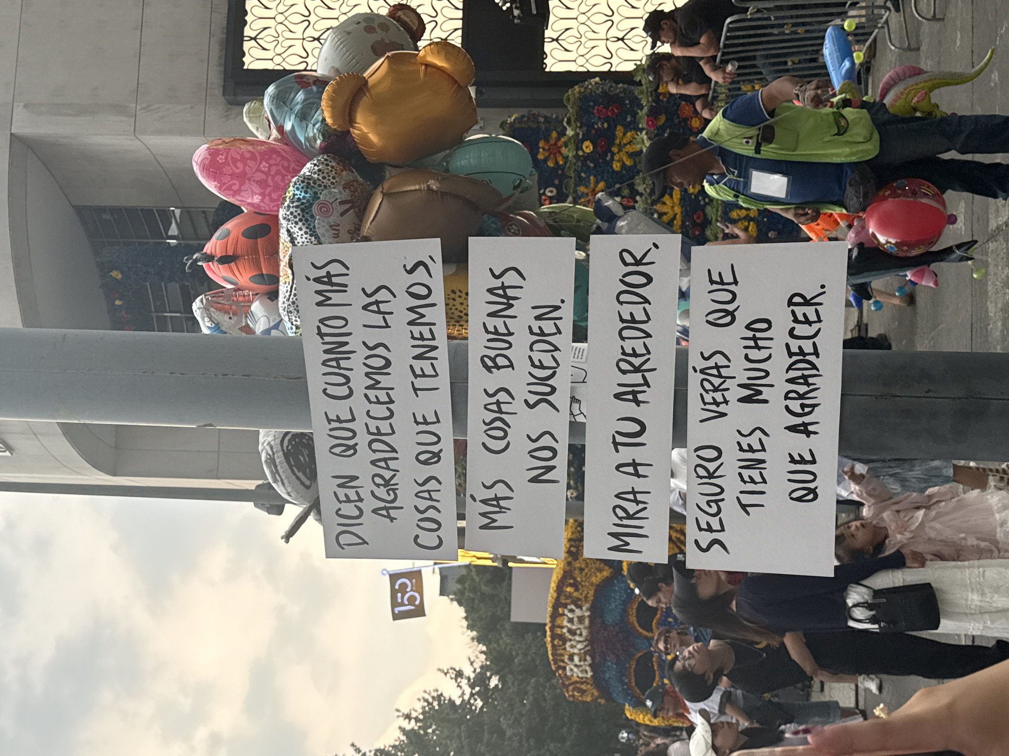 The image captures a scene in Mexico City, Mexico, centered around a pole adorned with balloons and signs. The signs, written in Spanish, contain phrases about gratitude and the positive effects of appreciation. A crowd of people is present, with some individuals visible around a floral arrangement. A person wearing a green vest and carrying a balloon stands amidst the crowd. The environment appears to be an urban setting, with buildings visible in the background.