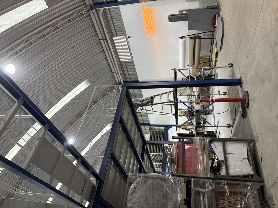 An industrial space with high ceilings, metal support beams, and corrugated metal roof. A person rests on a ladder connecting two levels of a metal structure. Another person works on equipment nearby. Packaging materials are stacked in the foreground. A machine with paper rolls stands against a wall. The space is illuminated by overhead lights and natural light through skylights. The scene is somewhat cluttered, indicative of ongoing operations.
