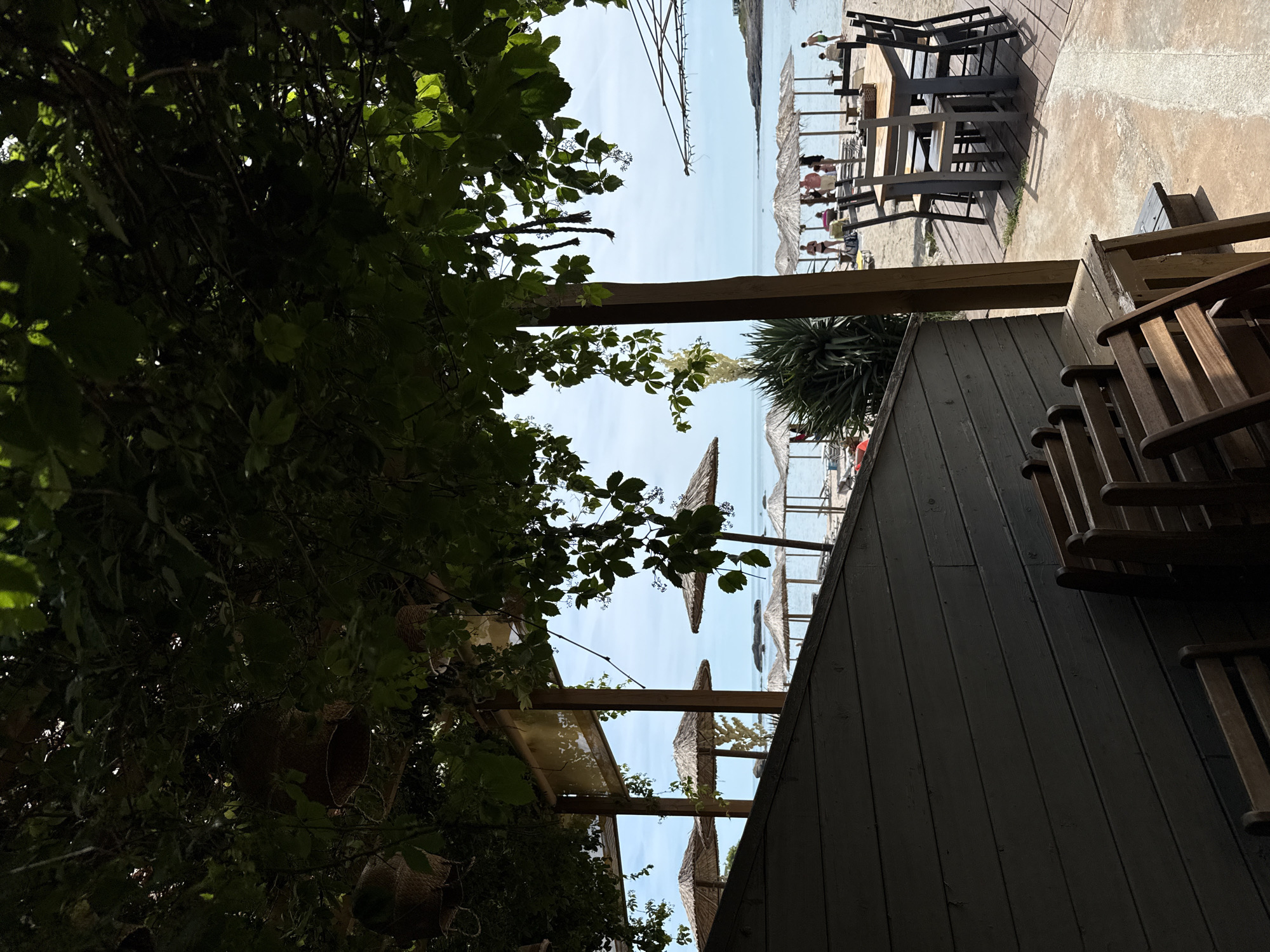 The image captures a view from a shaded outdoor seating area, looking out towards a beach on a sunny day. In the foreground, a dark wooden structure with horizontal planks partially obscures the view, with several empty wooden chairs stacked and arranged in front of it. Beyond this structure, the scene opens up to a sandy beach lined with straw umbrellas.
In the midground, several groups of people are visible on the beach, some standing near the umbrellas and others walking. The sea is calm and blue, with small waves lapping the shore. In the distance, the horizon is marked by a clear sky with some scattered clouds.
The overall impression is of a relaxed beachside establishment, likely a restaurant or cafe, offering a shaded respite from the sun with a view of the sea. The presence of tables and chairs suggests a place where patrons can dine or socialize. The scene is set in the city of Tsarevo, Bulgaria, known for its coastal resorts and beaches.