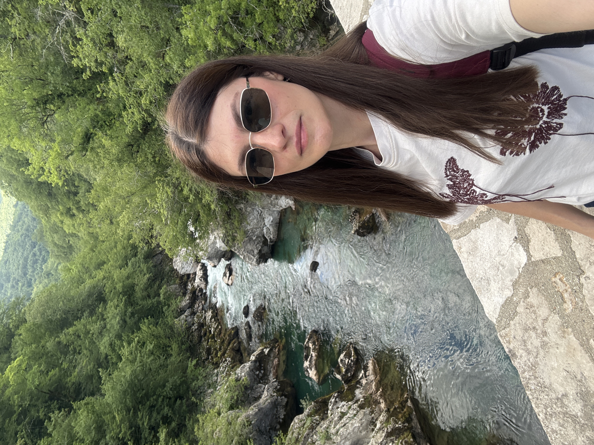 A woman is pictured in a medium shot, positioned on a stone bridge. Her long, brown hair flows over her left shoulder. She is wearing sunglasses and a white T-shirt with a brown floral design. A burgundy fanny pack is slung across her back. The background features a river flowing between rocky banks and lush green trees. The setting is identifiable as Međuriječje, Montenegro, based on the distinctive river and surrounding landscape.