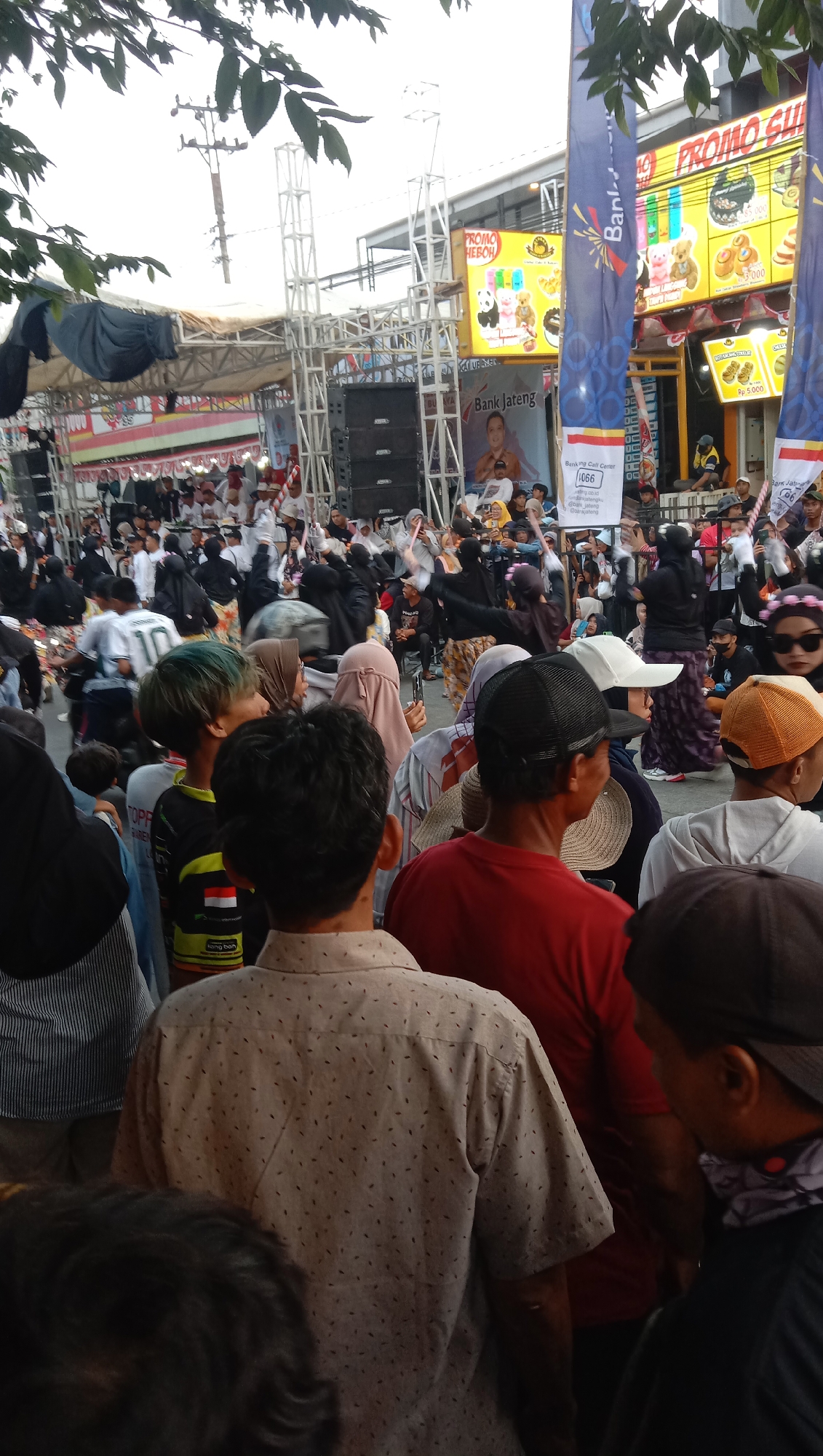 The image depicts an outdoor gathering in Jatisawit, Indonesia, likely a public event or market. A crowd of people is visible, many wearing casual clothing and some with religious head coverings. In the background, there are stages, sound systems, and large banners advertising promotions and a bank named "Bank Jateng". One banner prominently displays the words "PROMO GU" and "PROMO HEBOH" with images of teddy bears and pastries. The environment appears to be a street or open area, possibly a town square, with trees and utility poles also present. The overall atmosphere suggests a lively event with commercial activities.