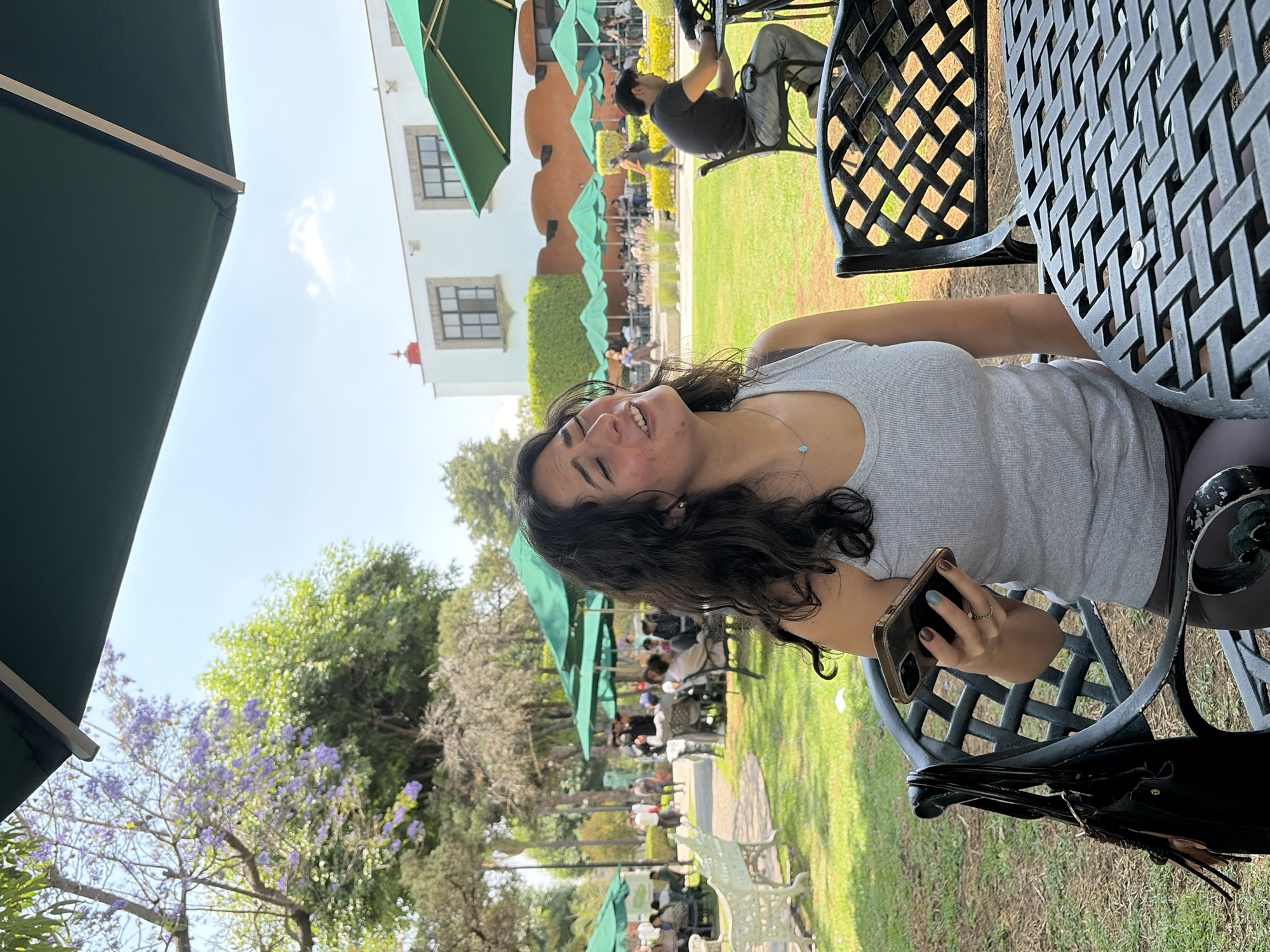 The image, rotated 90 degrees clockwise, captures a lively outdoor cafe or restaurant scene, likely in San Andrés Cholula, Mexico, under a bright, clear blue sky with wispy clouds. In the foreground, a young woman with long, dark, wavy hair is seated at a black metal outdoor table. She wears a grey ribbed tank top and a delicate necklace with a small turquoise pendant. Her head is tilted back as she laughs heartily, displaying a wide, joyful smile. She holds a smartphone with a gold case in her left hand; her nails show hints of light blue polish. A black bag hangs from her chair.
The setting is a spacious garden with lush green grass. Numerous green umbrellas are scattered across the scene, providing shade for other patrons seated at similar black metal tables and chairs in the midground and background, indicating a busy social gathering. A man in a black t-shirt and jeans is visible seated at a table further back. In the background stands a white, possibly colonial-style building with dark-framed windows and a distinct reddish architectural detail on its roof. Various trees, including one adorned with vibrant purple blossoms (likely Jacaranda), enhance the scenic garden, alongside white park benches. The overall atmosphere is cheerful and relaxed, depicting people enjoying a pleasant daytime outing. No discernible text is visible in the image.