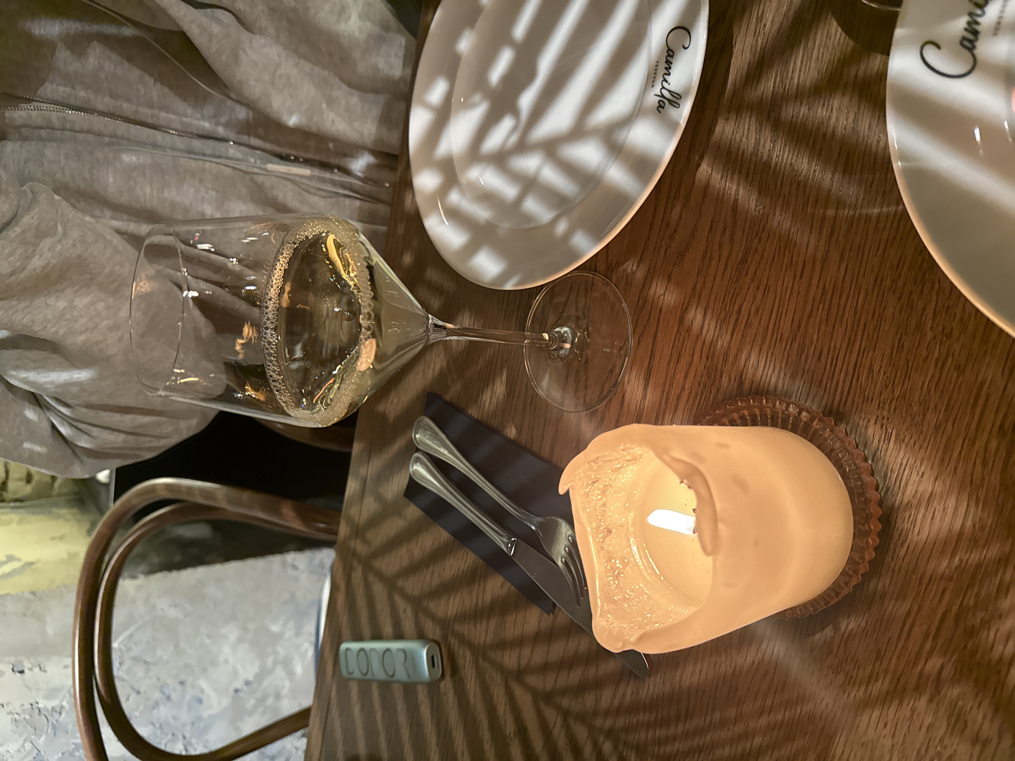 The image shows a table setting at Camilla Yerevan, located in Yerevan, Armenia. The table is made of dark-stained wood and has a white plate with the restaurant's name on it. A wine glass filled with a light-colored beverage lies on its side. There is also a lit candle in a glass holder and silverware resting on a black napkin. A chair with a metal frame is partially visible.