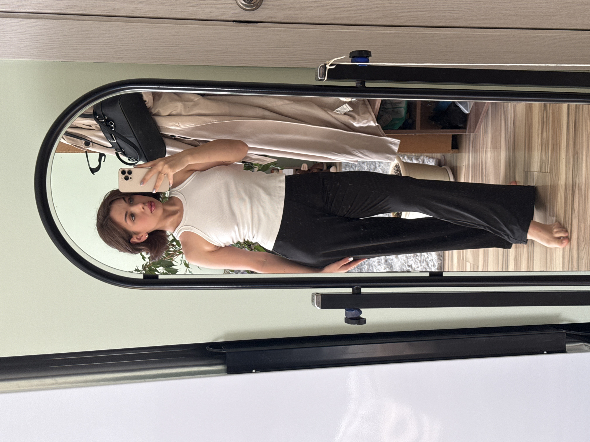A person is seen in the reflection of an oval-shaped mirror. The person has shoulder-length brown hair and is wearing a white tank top and black pants. They are holding a smartphone up to take a selfie. In the background, there is a partially visible room with a shelf, clothes, and a plant. The floor has a wood-like texture. The mirror is hung on a light green wall. The bottom of the image shows a white surface. It's impossible to ascertain the location as Sarapul, Russia.