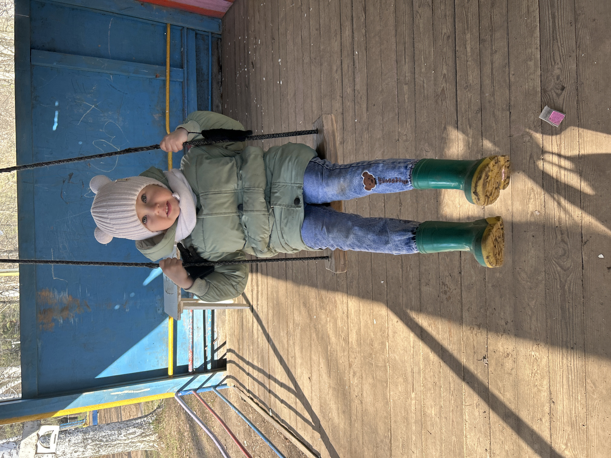 A young child sits on a wooden swing, looking directly at the camera. The child wears a light green puffy jacket, blue jeans with a brown teddy bear patch on the right thigh, bright green rubber boots with yellowed soles, a white knit hat with ear-like details, and a white scarf. Their hands grip the dark ropes of the swing.
The swing is suspended over a rustic wooden plank floor, which shows signs of wear and tear. To the child's left is a blue painted wooden wall or structure, covered in white scratches and graffiti. To their right and in front of them, colorful metal railings (red, blue, yellow, green) are visible, leading down. Beyond the railings and structure, trees and glimpses of ground are in the background, suggesting an outdoor setting in a playground.
Strong natural sunlight illuminates the scene, casting distinct, long shadows across the wooden floor, indicating a sunny day. A small, square, pink and white object, possibly chalk or a small toy, is visible on the wooden floor to the right of the child's feet. The text on the brown patch on the child's jeans appears to say "Flower Kid" beneath the teddy bear graphic. The child's expression is neutral, possibly a slight smile. This scene is set in Chaikovsky, Russia.