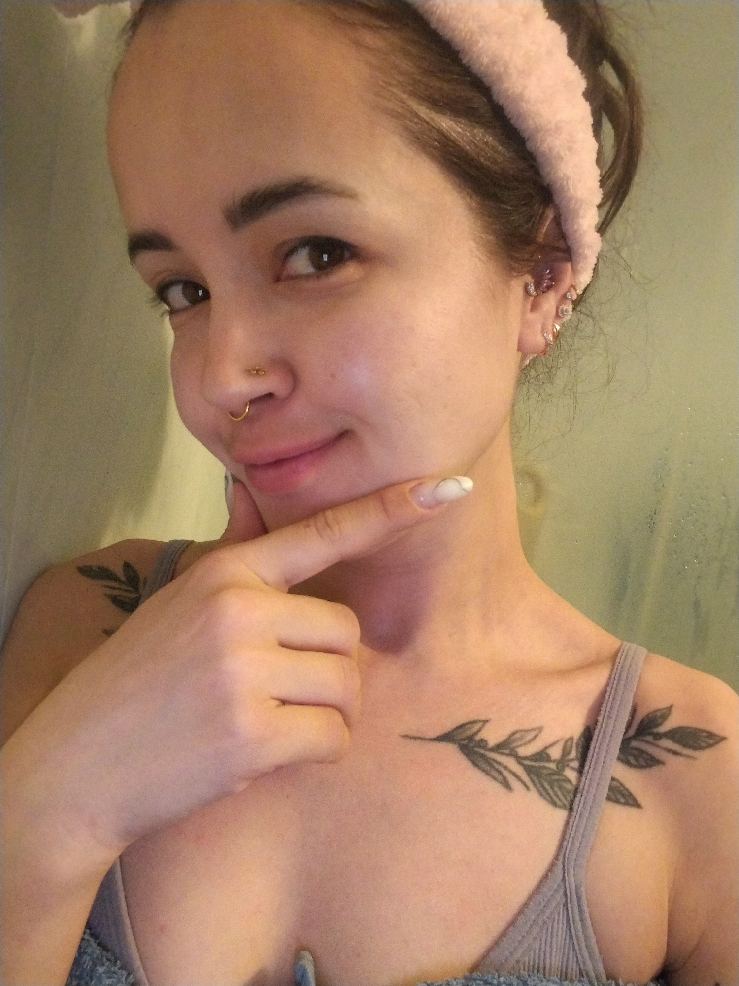 A woman is posing for a selfie. She has brown hair pulled back with a pink headband. She has multiple piercings in her left ear and a gold ring in her nose. She's wearing a gray tank top and has two leaf-branch tattoos, one on her left shoulder and one on her chest. Her right index finger is pointing at her chin. The background appears to be a blurred wall, possibly in a bathroom, with condensation visible. There is no information related to Noviy, Russia, in the image.