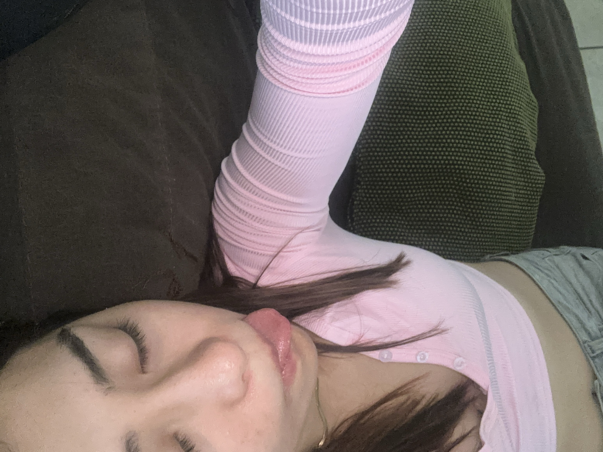 A person is lying down, seemingly on a sofa, with their eyes closed and their tongue sticking out. They are wearing a pink, long-sleeved, ribbed shirt and grey bottoms. The sofa has two sections, one olive green and the other dark brown. There is no indication that this image was taken in the city of Puebla, Mexico.