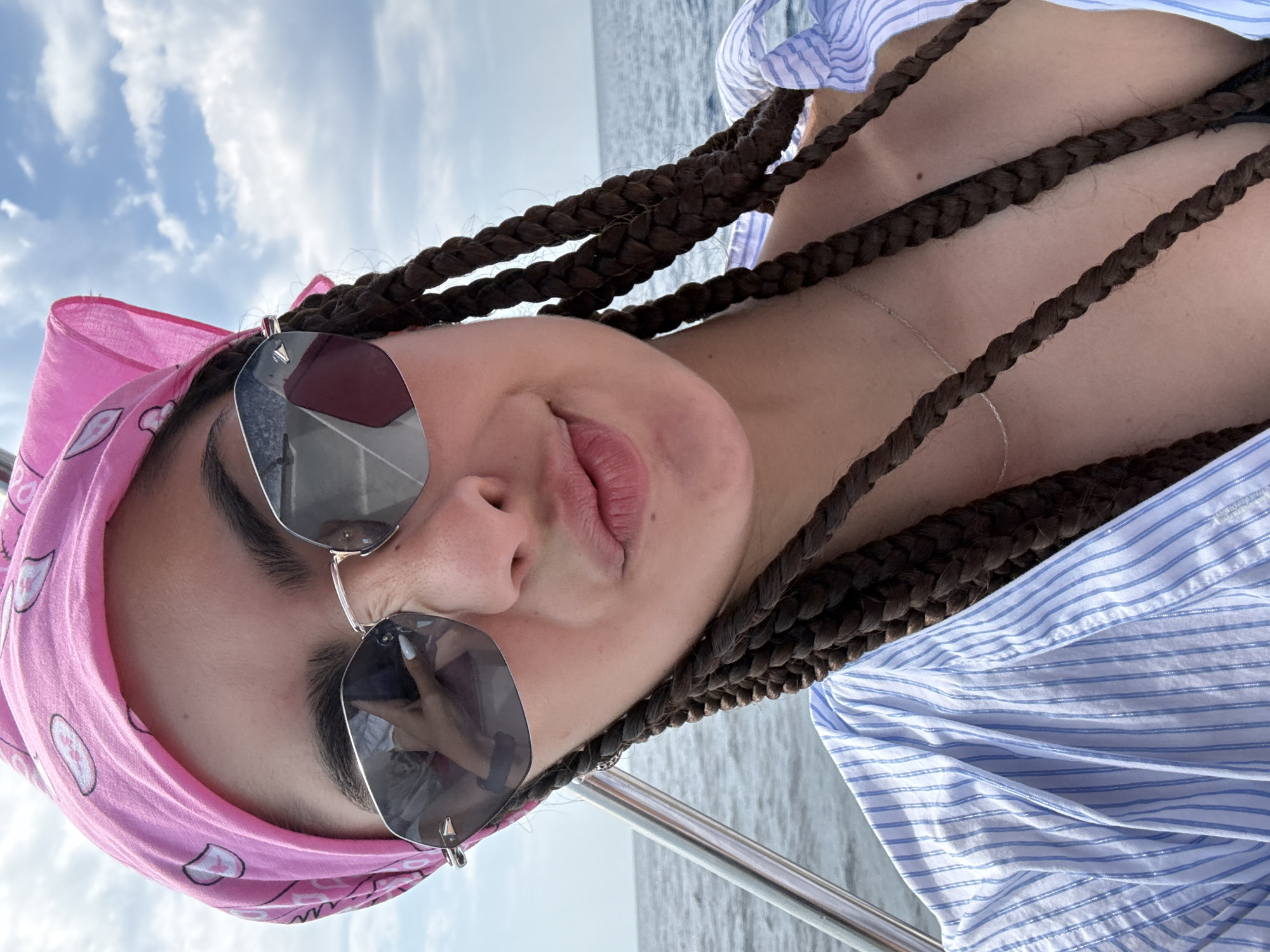 The image is a selfie of a young woman with dark braided hair, wearing a pink bandana, sunglasses, and a light blue and white striped shirt. She is outdoors, on a body of water, likely a sea or ocean, with a cloudy sky in the background. The woman is looking towards the camera with her lips puckered. A metal railing is visible on the left side of the image. The overall scene suggests a leisurely day on the water. The location is described as unknown, Greece.