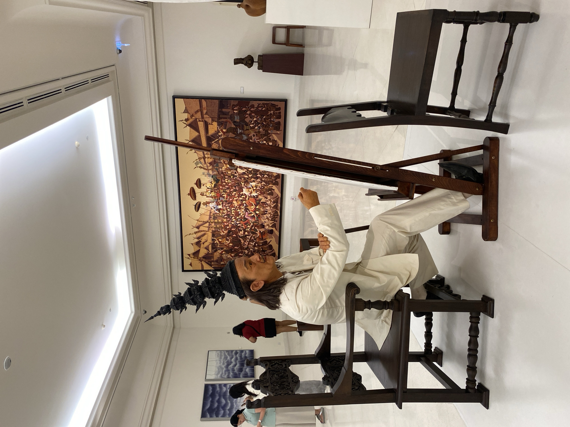 A statue of an artist in a white suit and black, spiked headdress sits at an easel in what appears to be a gallery in Bangkok, Thailand. He appears to be painting an unseen canvas. Behind him is a large painting depicting a historic Thai scene. Other chairs and art pieces are in the background, and a few other people can be seen in the distance. The gallery is brightly lit with white walls and appears to be modern.