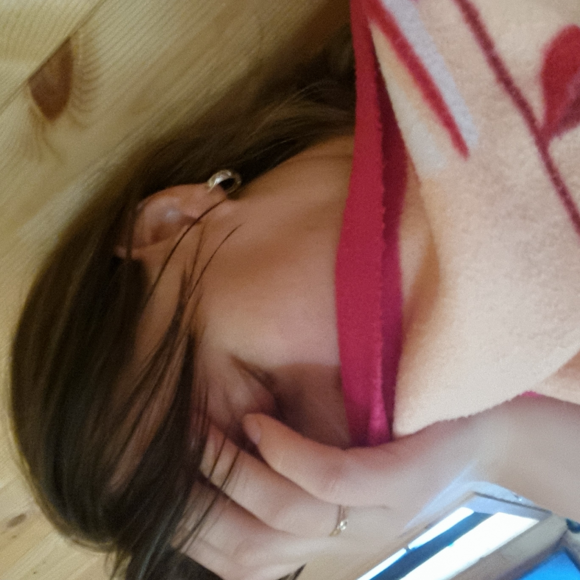 A close-up, indoor, medium shot shows a person's face partially obscured by their hand and a pink and beige blanket. The person has dark brown hair and is wearing a small hoop earring on their visible ear. The background consists of light-colored wooden planks. The overall scene is dimly lit, with a shallow depth of field. The image appears to be taken from a low angle.