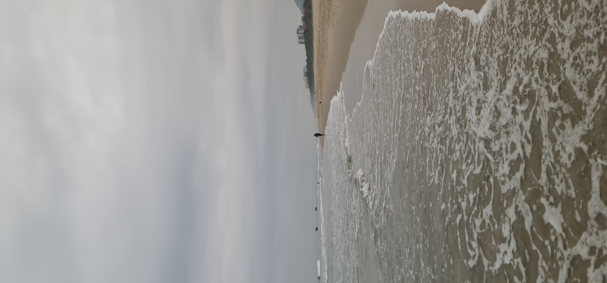 This image captures a wide-angle view of a beach scene on an overcast day. The vast, light grey, cloudy sky dominates the upper half of the frame, suggesting subdued lighting and possibly cool weather. Below the sky, a wide expanse of light brown sand stretches horizontally, forming the beach.
To the right, the ocean waves are actively breaking on the shore, creating a foamy white pattern that spreads across the wet sand. The water appears a muted greyish-brown, reflecting the cloudy sky.
Several people are visible along the waterline. A solitary figure, dressed in dark clothing, is walking along the edge of the incoming waves in the mid-ground, heading towards the left. Further in the distance, along the receding shoreline, other much smaller and indistinct figures can be seen, also near the water.
In the far background, along the horizon where the beach meets the land, a line of buildings (possibly hotels or apartment complexes) can be discerned, partially obscured by the haze, with faint outlines of hills or mountains rising behind them. The overall impression is one of calm and peace, characteristic of a quiet beach day. Given the location context, this scene depicts a beach in Da Nang, Vietnam. There is no visible text.