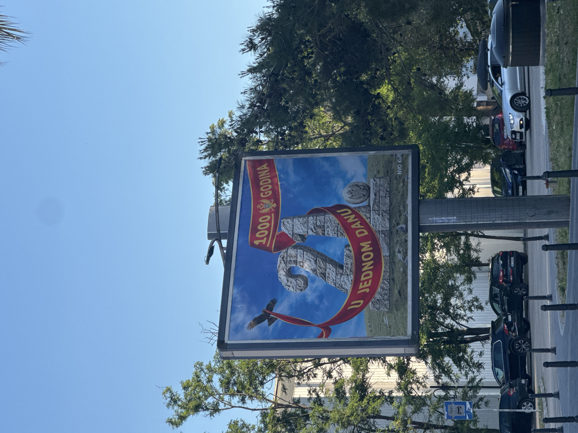 The image depicts a billboard in Bar, Montenegro, set against a blue sky with scattered clouds, framed by trees. The billboard displays a design commemorating "1000 godina" (1000 years), featuring a large stone number "2" with a red ribbon wrapping around it that reads "U jednom danu" (In one day), and an image of a double-headed eagle. The billboard is supported by a gray metal pole with street signs. The background shows a street with parked cars, a sidewalk, and bollards. The image is taken at a slight upward angle.