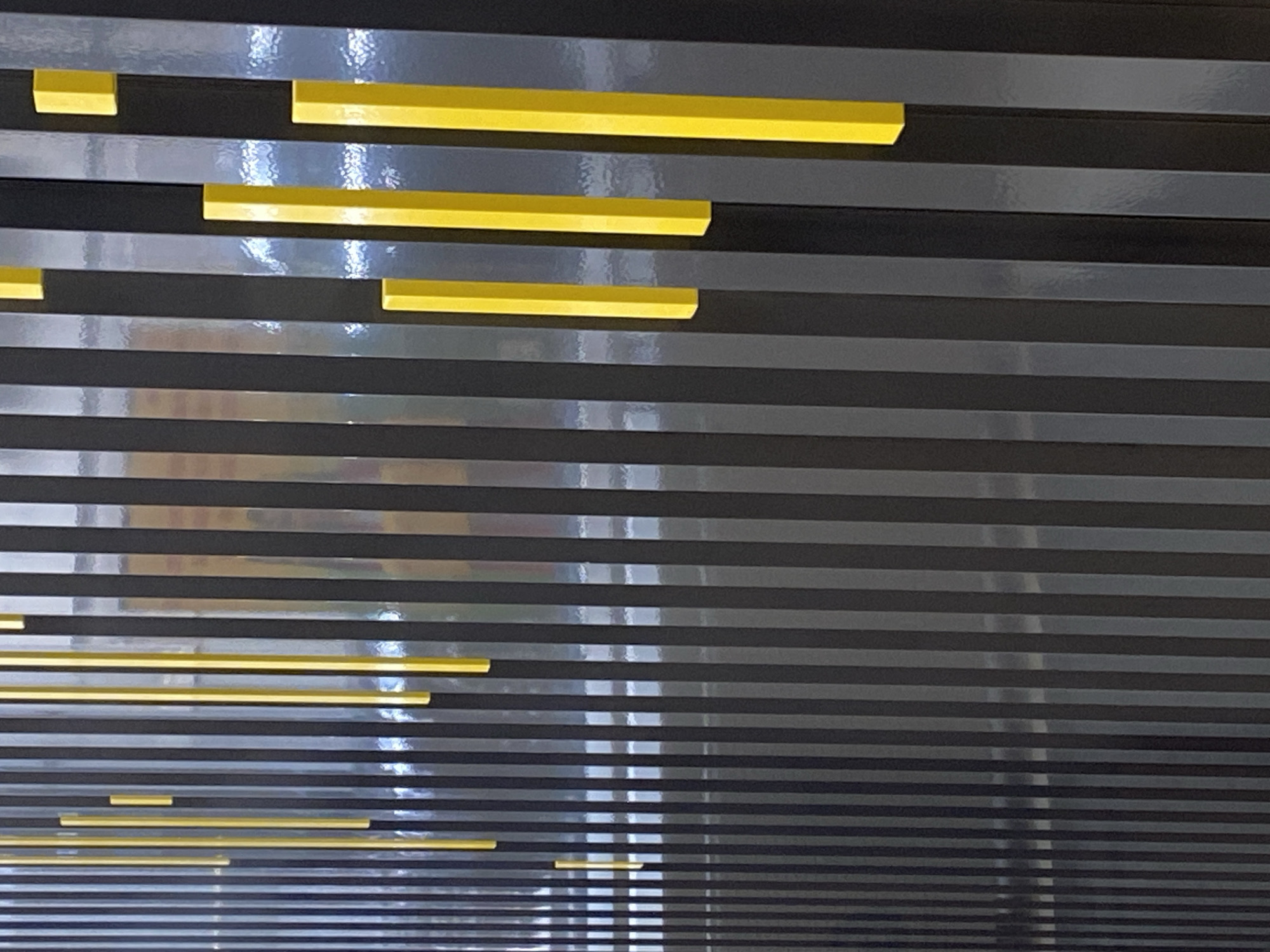 The image captures a modern, decorative wall or paneling featuring horizontal stripes in dark grey and bright yellow. The background comprises numerous parallel dark grey or black stripes, many of which exhibit a highly glossy, reflective surface. These reflective surfaces show vertical streaks of reflected light, suggesting illumination from overhead light sources. Layered unevenly across these dark stripes are several rectangular bars of vibrant yellow. These yellow elements vary in length and appear to be slightly raised or protruding from the surface, adding a three-dimensional quality to the design. The composition is abstract and static, with no people, discernible activities, or visible text. The stark contrast between the glossy dark background and the matte, bright yellow accents creates a contemporary aesthetic. The setting is an interior space, possibly located in Sofia, Bulgaria, as provided by the context.