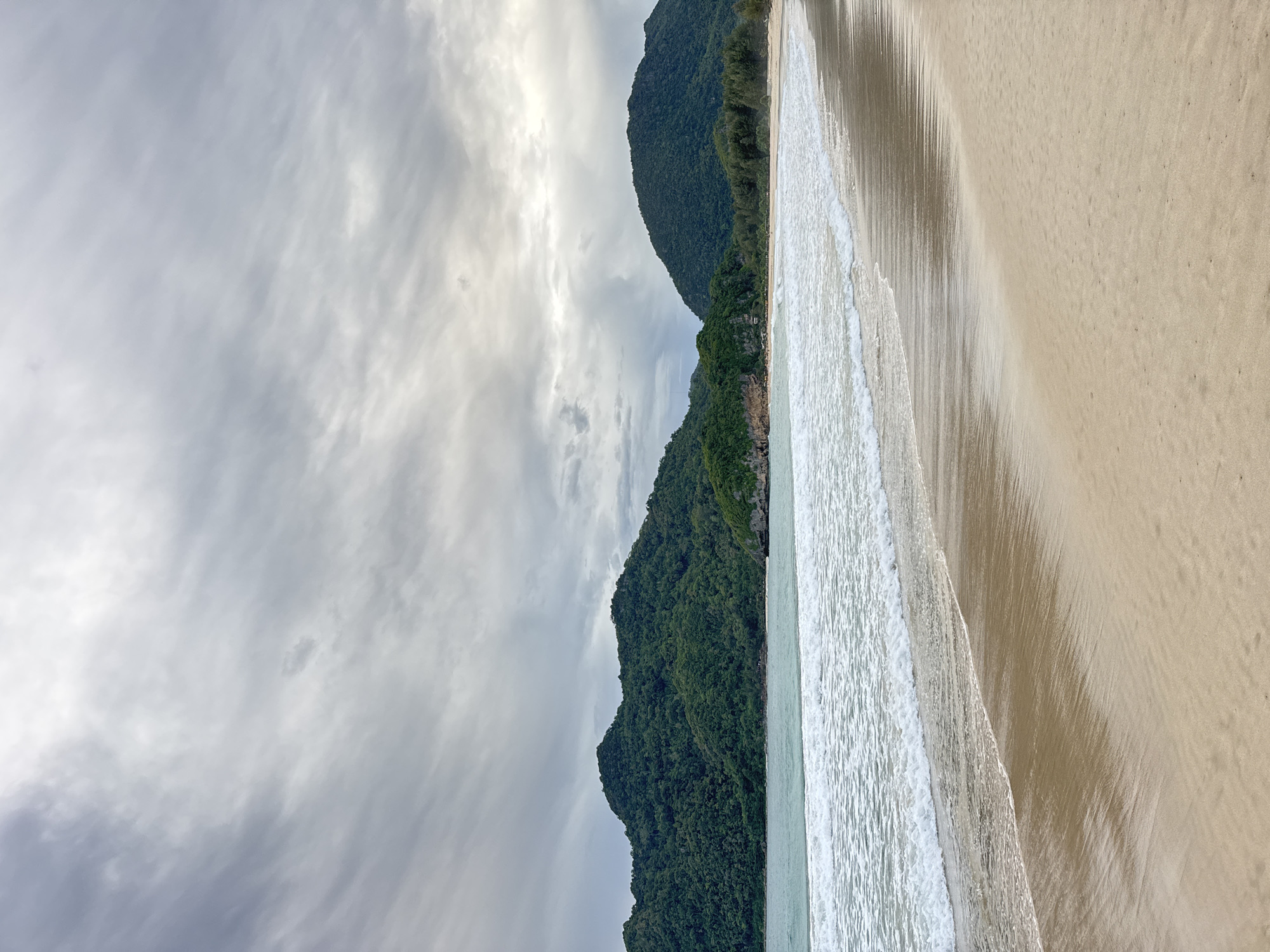The image depicts a serene beach scene with a backdrop of lush, green mountains. The sky is overcast with a heavy, textured layer of grey clouds, hinting at potential impending rain or a recent storm. The ocean is calm, with gentle white-capped waves lapping against the shore. The sand is a light, warm tan, showing subtle patterns from the receding tide. In the distance, the mountains are densely covered in tropical foliage, with some rocky outcrops visible along the coastline. The overall atmosphere is tranquil and natural, capturing the essence of a coastal landscape. This scene is located in Meunasahbale, Indonesia, a coastal area known for its natural beauty and verdant mountainous terrain meeting the sea. No people or specific objects are discernible in the image, emphasizing the unspoiled nature of the environment.