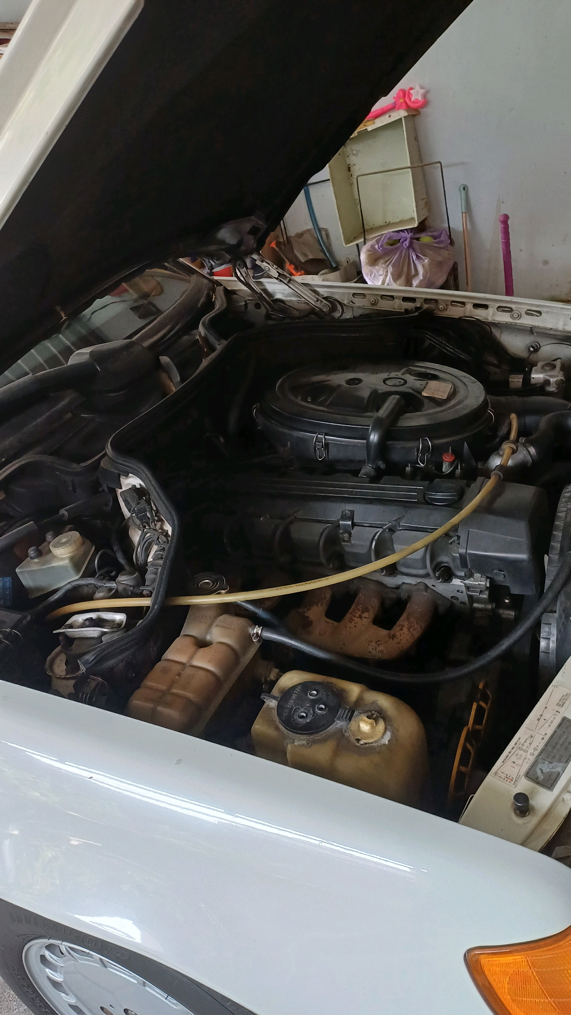 The image shows the engine bay of a white car. The hood is open, revealing the engine and various components. There is a large black air filter housing on top of the engine, with some hoses and wires connected to it. To the left of the engine, there is a clear plastic coolant reservoir with markings. To the right, a rusty exhaust manifold is visible. The car appears to be parked indoors, possibly in a garage or workshop, as indicated by the presence of a wall with some storage items like bags and cleaning tools. The lighting suggests an indoor environment. The image is taken from a slightly elevated perspective, looking down into the engine bay. The overall impression is that of a car undergoing maintenance or inspection. This scene is located in Karangwuni, Indonesia.