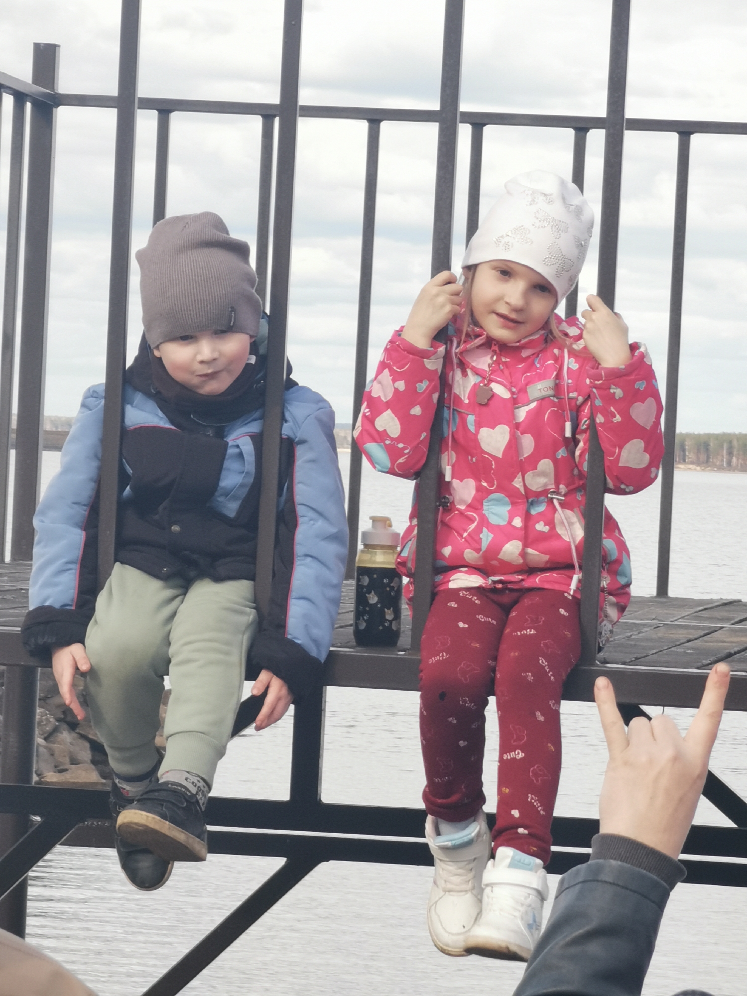 Two children, a boy and a girl, are sitting on a metal structure that resembles a swing. The boy on the left wears a grey knit hat, a blue and black jacket, green pants, and black sneakers. The girl on the right wears a white hat with butterfly patterns, a pink jacket with white heart patterns, and red pants with white lettering. A water bottle sits next to the girl. A person's hand in the lower right corner of the frame makes a "rock on" hand gesture. In the background, a body of water stretches to the horizon under a cloudy sky.