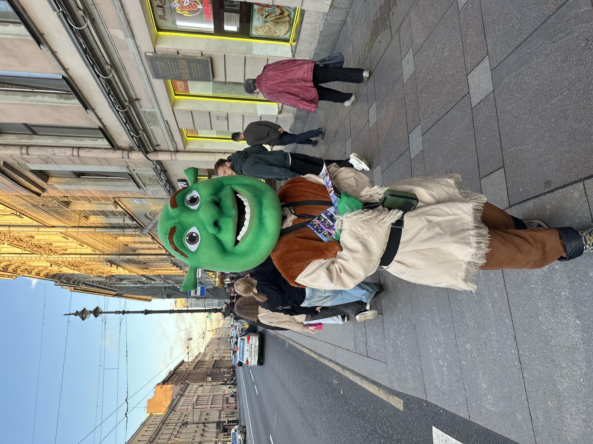 A person in a Shrek costume stands on a sidewalk in Saint Petersburg, Russia, holding brochures. The character is wearing a full green Shrek head, a tan tunic with a brown vest, and brown pants. The Shrek costume wearer is facing the camera, with other pedestrians walking in a line behind them on the sidewalk. A street with cars and buildings lines the left side of the image. The buildings appear to be historic and have ornate architectural details. The sky is clear and blue, and the sun is casting a warm light on the buildings. The sidewalk is paved with gray tiles.