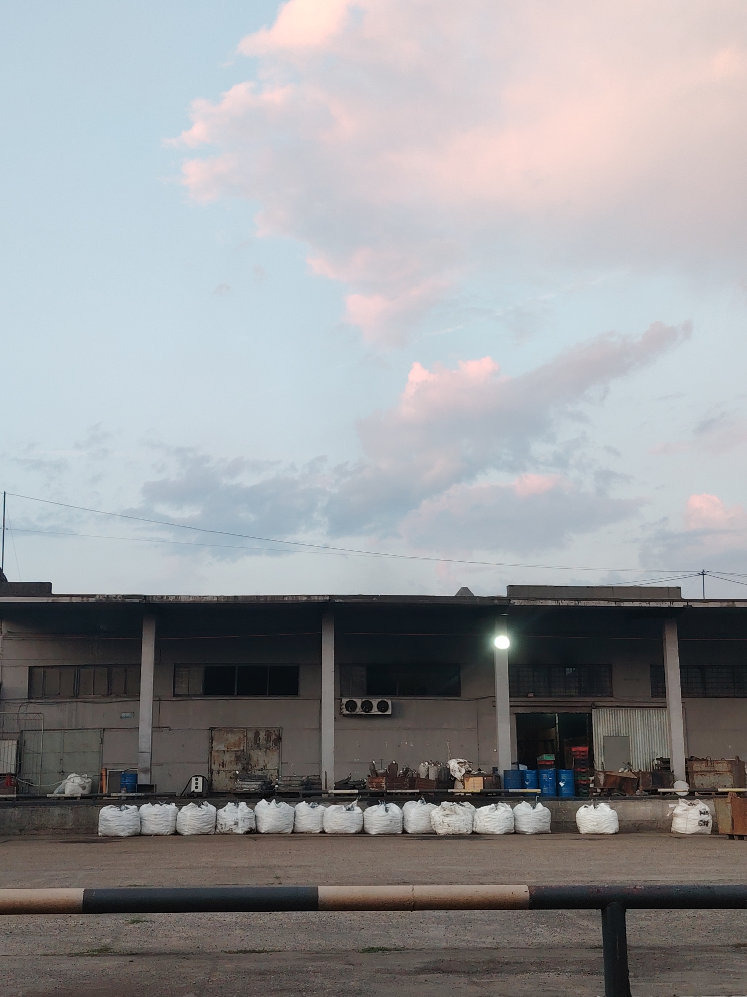 The image depicts an outdoor industrial scene in Noviy, Russia, under a pale sky with soft pink and white clouds. In the foreground, a dark metal railing with a brown and black striped top runs horizontally. Behind the railing is a paved surface. The middle ground features a row of large, white, opaque sacks lined up on a concrete loading dock. To the left of the sacks, a blue drum is visible. The background shows the facade of a long, grey industrial building with multiple windows, some of which appear to be boarded up or darkened. A bright light source illuminates the area near a doorway on the right side of the building. Various objects, including barrels, crates, and metal structures, are scattered around the loading dock and visible through the open doorway. An air conditioning unit is mounted on the exterior wall of the building. The overall impression is that of a storage or logistics facility.