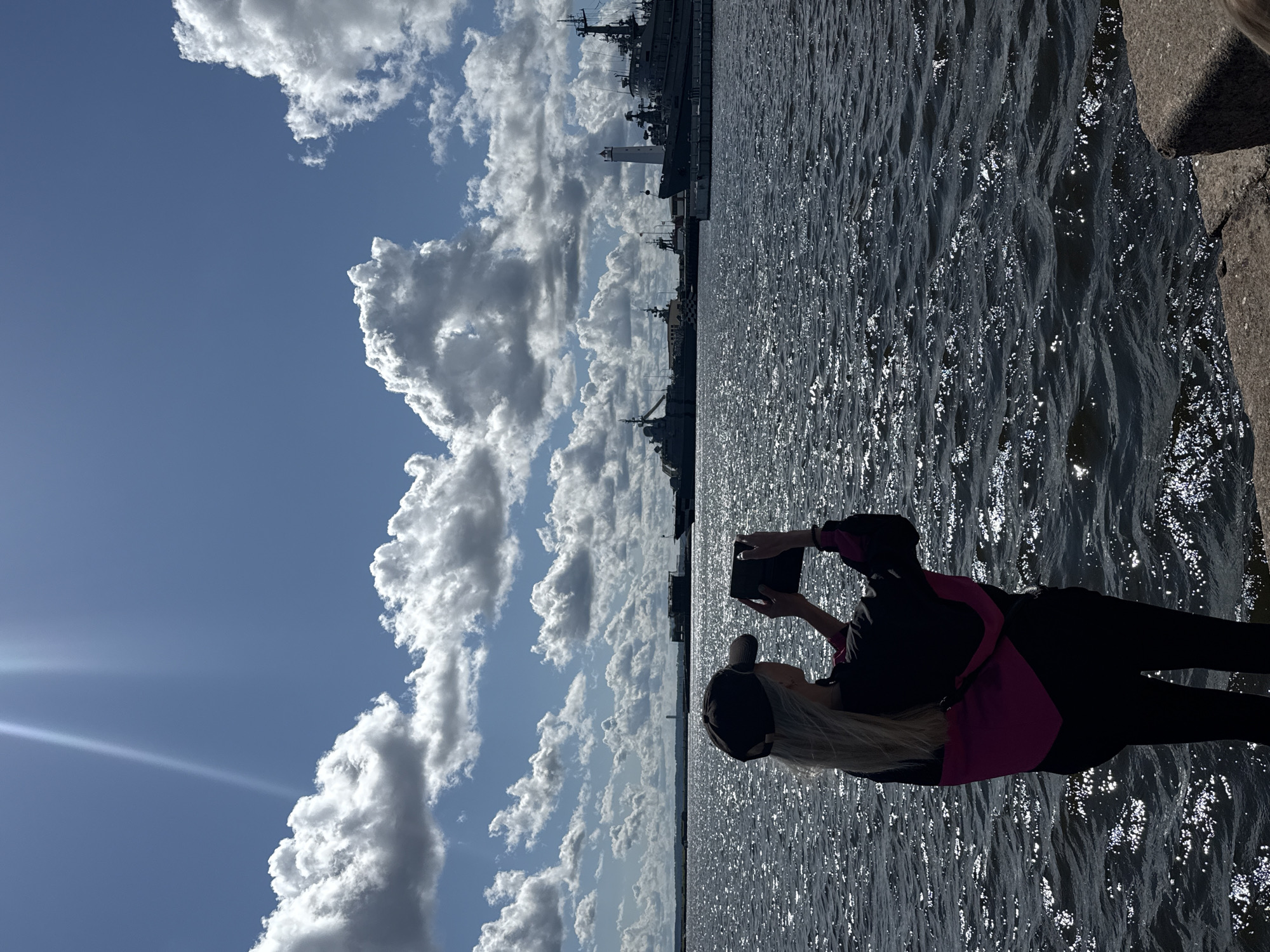 The image depicts a woman wearing a black baseball cap and a black and pink jacket, standing on a stone embankment overlooking a body of water. She is holding a tablet in her hands, appearing to take a photograph or video of the scene in front of her. The water is dark and choppy, reflecting the sunlight.
In the background, several large ships, including what appears to be a lighthouse, are docked at a port. The sky is a bright blue with scattered white cumulus clouds. The scene suggests a sunny day in an urban port environment.
The location is the city of Kronstadt, Russia, a naval fortress town located on Kotlin Island at the entrance to the Gulf of Finland. The presence of the ships and the port infrastructure strongly supports this identification. The woman is positioned in the foreground, facing away from the camera, emphasizing the vastness of the sea and the maritime activity.