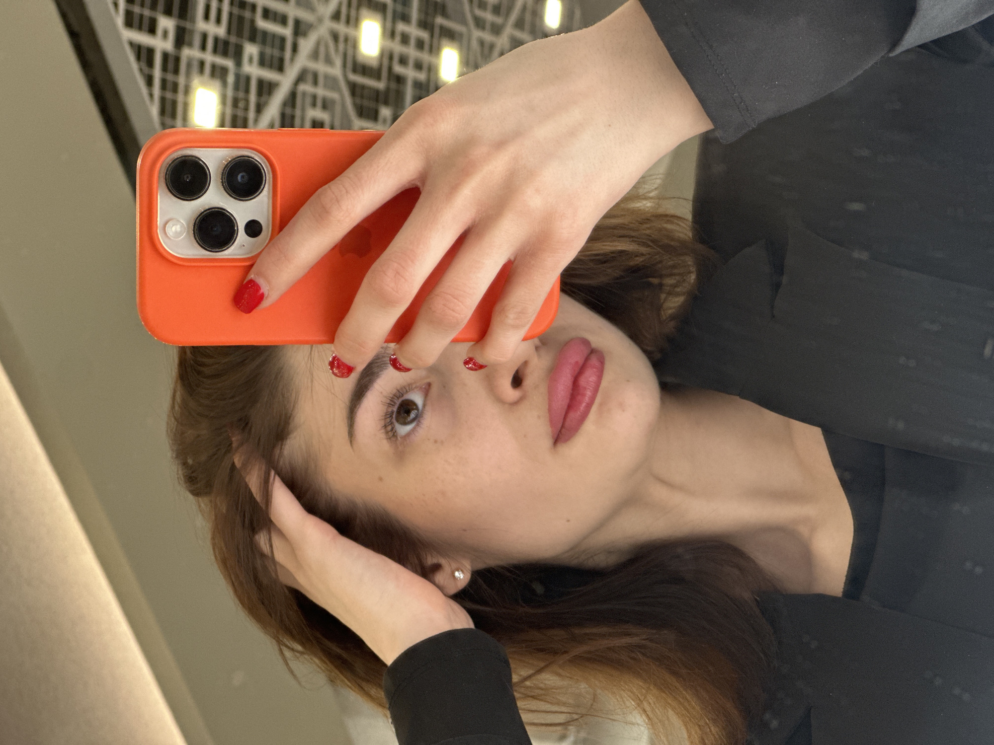 A female individual is depicted in a mirror selfie within an indoor setting in Yekaterinburg, Russia. The subject is a woman with brown hair, fair skin, noticeable freckles on her nose and right cheek, and pink-lipsticked lips. Her visible right eye, with a brown iris and long eyelashes, gazes slightly upward. She wears a black long-sleeved top under a black blazer. A small, circular silver stud earring is visible in her right earlobe. Her left hand is raised, with fingers partially intertwined in her hair on the left side of her head. Her right hand holds an orange-cased smartphone, possibly an iPhone due to the three-lens camera array visible on the back, obscuring the left side of her face. The fingernails on both visible hands are painted red. The background, reflected in the mirror, shows a light-colored ceiling with a grid pattern incorporating recessed square light fixtures and a linear light strip, suggesting a modern commercial or public interior. The exact location within Yekaterinburg is not discernible from the available visual information.