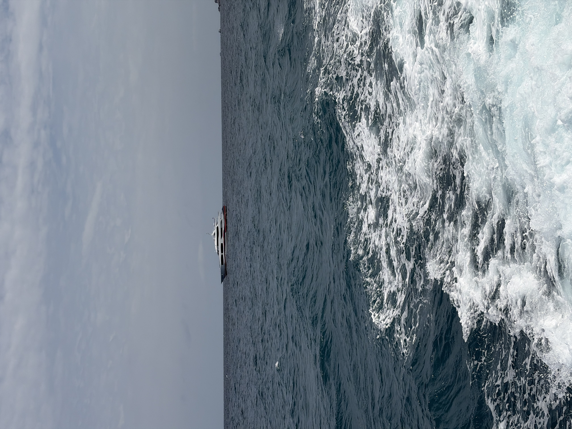 An image taken from a boat in the ocean off the coast of Ko Pha-ngan District, Thailand, shows a white and red yacht in the distance. The water is dark blue with whitecaps and churned up wake in the foreground, indicating movement. The sky is overcast with a light gray hue.