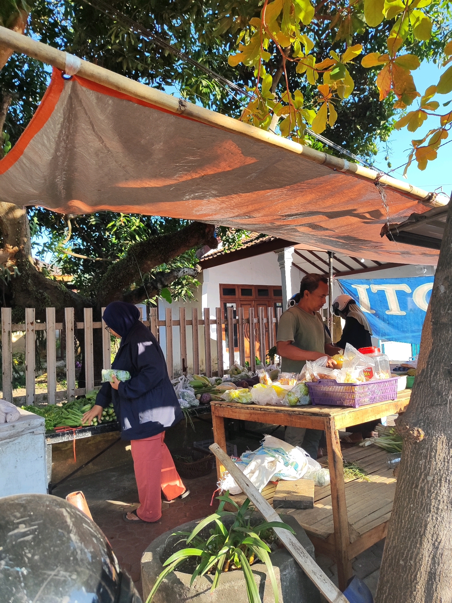 This image depicts an outdoor market scene in Bondowoso, Indonesia, on a sunny day. A makeshift stall is set up under a tarp supported by bamboo poles. The tarp, colored orange and beige, provides shade for the vendors and their goods. In the foreground, a woman wearing a dark blue hijab and jacket, along with coral-colored pants, is seen examining produce. She is holding a bunch of green vegetables in her left hand. In the mid-ground, a man in a green t-shirt stands behind a table laden with various items, including plastic bags of produce, a purple basket containing yellow items, and jars. He appears to be interacting with a person partially visible behind him, who is wearing a helmet. The background reveals a white wooden fence, trees, and a building with a blue banner displaying white lettering that appears to be "ITO". The ground is paved with bricks. To the left, part of a black motorcycle is visible. The overall atmosphere suggests a typical local market with vendors and customers engaged in commerce.