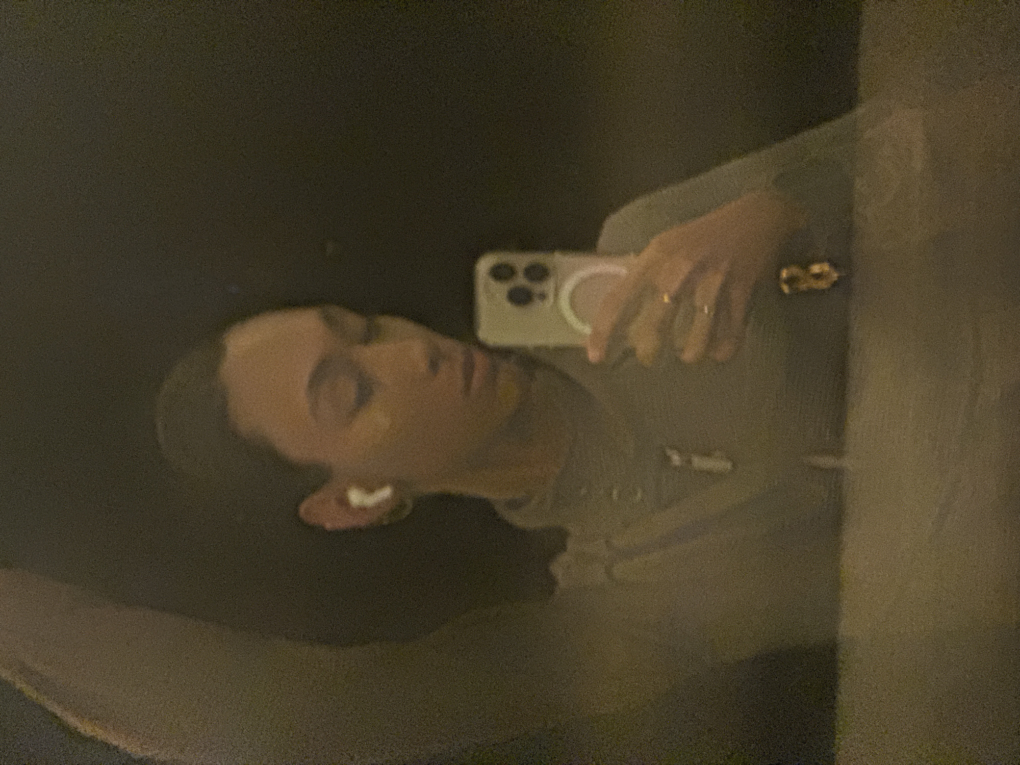 The image shows a person taking a selfie in a mirror. The person is holding an iPhone. They appear to be wearing a sweatshirt. There is an AirPod in their ear. The setting is dimly lit, with dark background and limited visibility. The location cannot be determined from the image.