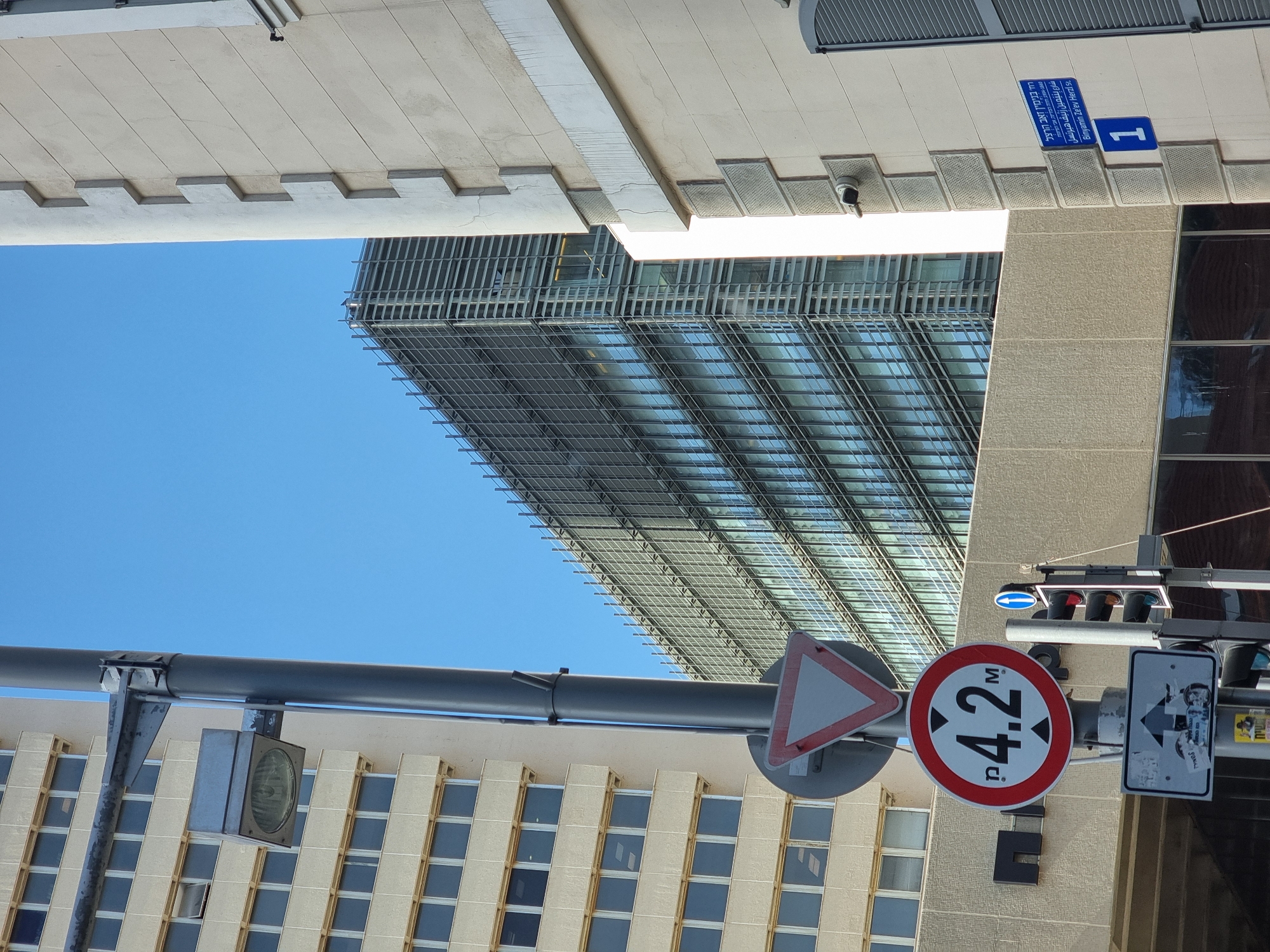 The image shows a low-angle view in an urban setting, specifically Tel Aviv, Israel, as indicated by the street sign. The sky is clear and blue. A modern skyscraper with a grid-like facade dominates the central background. Buildings on either side of the frame partially obscure the view upwards, creating a sense of enclosure. A pole with traffic signals and signage is prominent in the foreground. The signage includes a triangular yield sign and a circular height restriction sign displaying "4.2M" in bold numerals. The street sign is blue and white, with "Binyamin Ze'ev Herzl St." written in Hebrew, Arabic, and English, and the number "1". The overall impression is of a modern cityscape with visible infrastructure and signage.