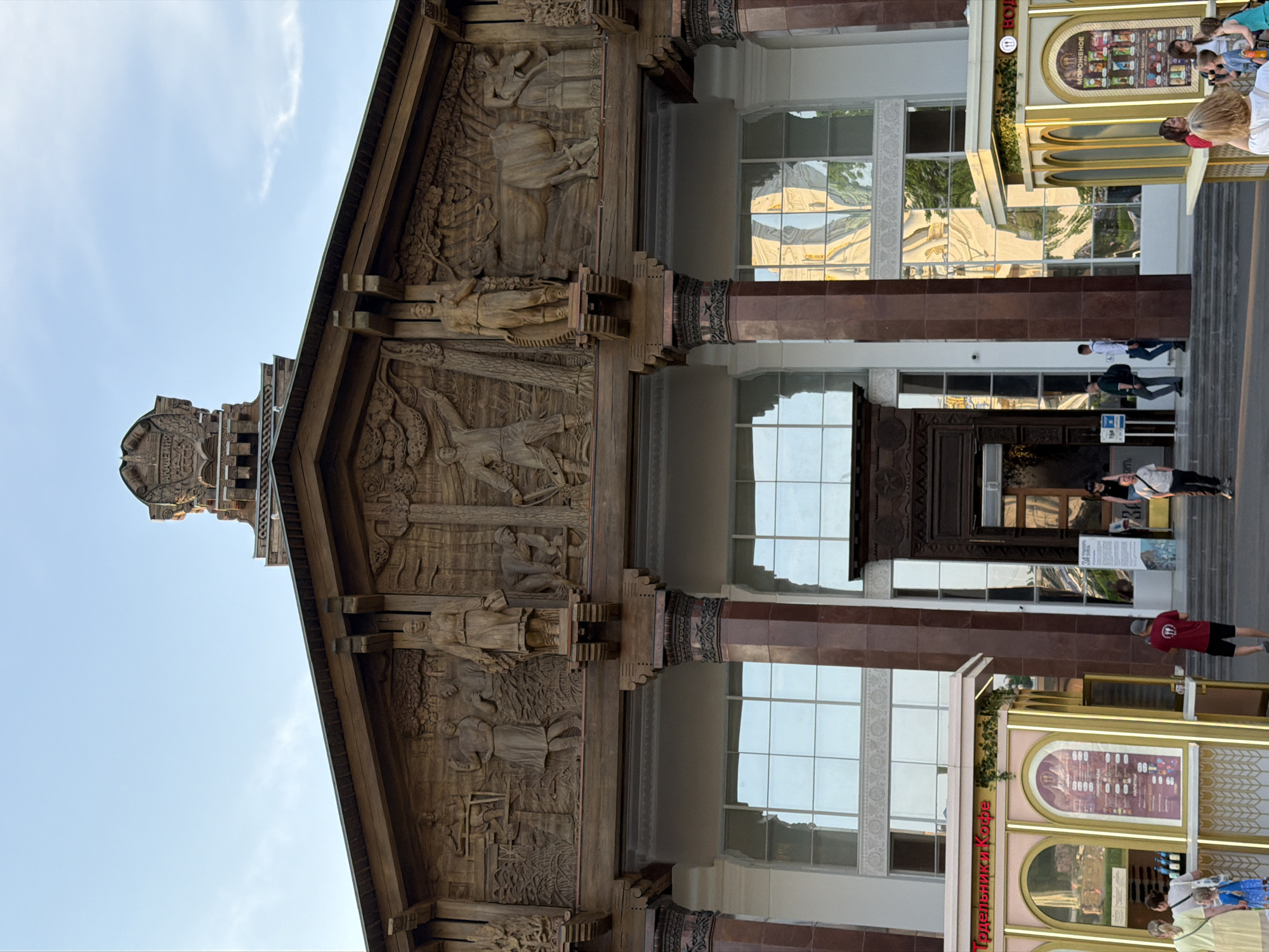 The image captures a frontal view of the facade of the Pavilion No. 58 "Agriculture" at VDNKh (Exhibition of Achievements of National Economy) in Moscow, Russia. The building features ornate wooden carvings depicting scenes of agricultural life. The facade consists of a central section with tall windows reflecting the sky and nearby structures. To the sides, there are brown tiled pillars and decorative elements. People are visible walking and standing in front of the building, including one wearing a red shirt. A bright, golden-toned structure, likely a concession stand or shop, is positioned to the side. The sky is clear and blue, and the overall setting suggests a sunny day.