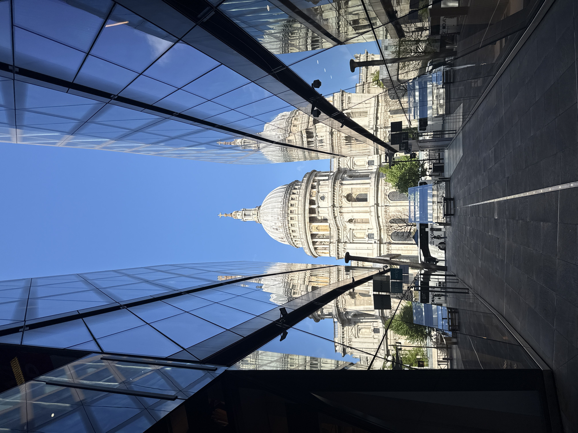 The image captures St. Paul's Cathedral in London, United Kingdom, framed by modern glass buildings. The cathedral dominates the view, seen between the converging lines of the adjacent buildings. The glass surfaces of the buildings reflect the cathedral, creating a mirrored effect and adding to the complexity of the visual composition. The sky is clear and blue. The architecture combines the historic cathedral with contemporary structures. The foreground consists of a narrow paved walkway with benches.