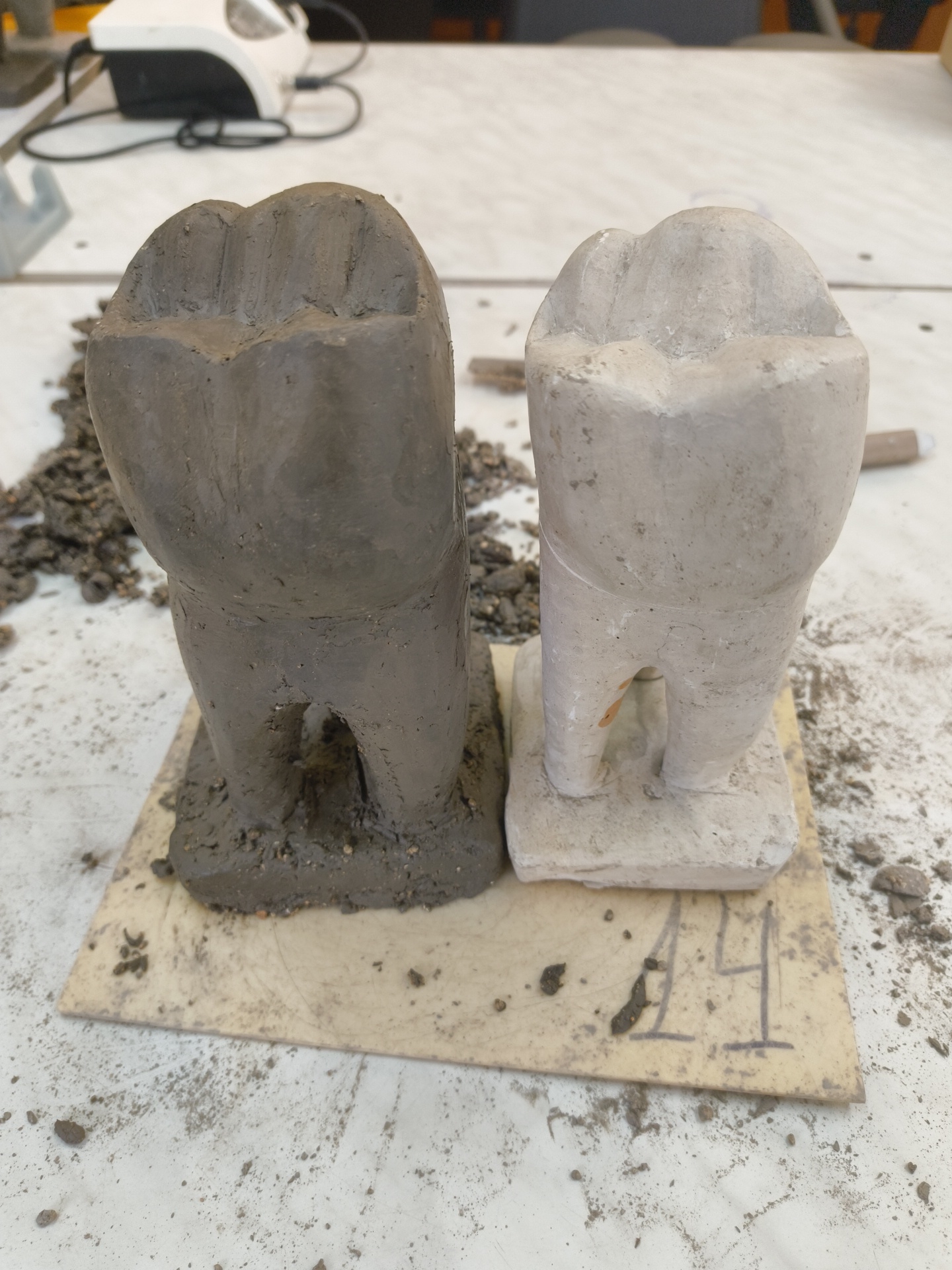 The image shows two tooth-shaped sculptures, one dark gray and the other light gray, standing side-by-side on a rectangular, light-colored tile with the number "44" written on it. The sculptures appear to be made of clay or concrete. The scene is set on a cluttered white table with debris scattered around. In the background, a white object with a power cord is visible. The city of Sofia, Bulgaria, cannot be confirmed as the location based on the visual information provided.