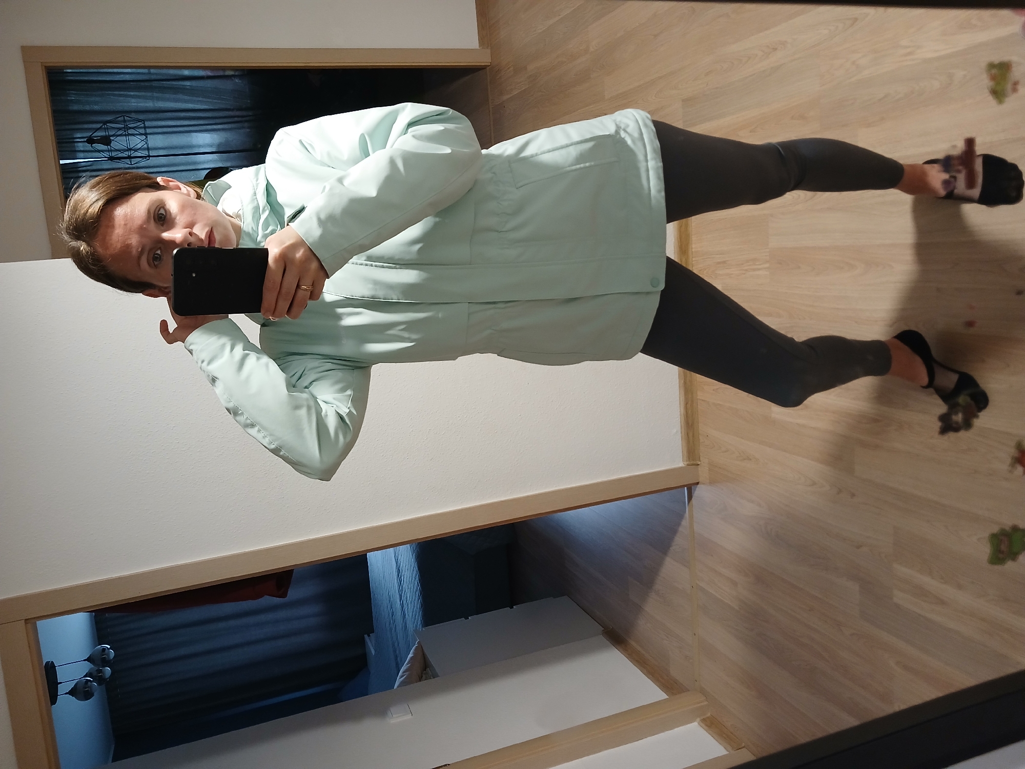 A person is standing in a room, taking a mirror selfie with a smartphone. The person is wearing a light mint green jacket and dark leggings. They are looking at the camera and adjusting their hair. The room has light wood floors and white walls. There is a doorway in the background leading to another room with a bed and curtains. The scene appears to be a residential interior. There is no specific information to determine the location in the city of Yekaterinburg, Russia.