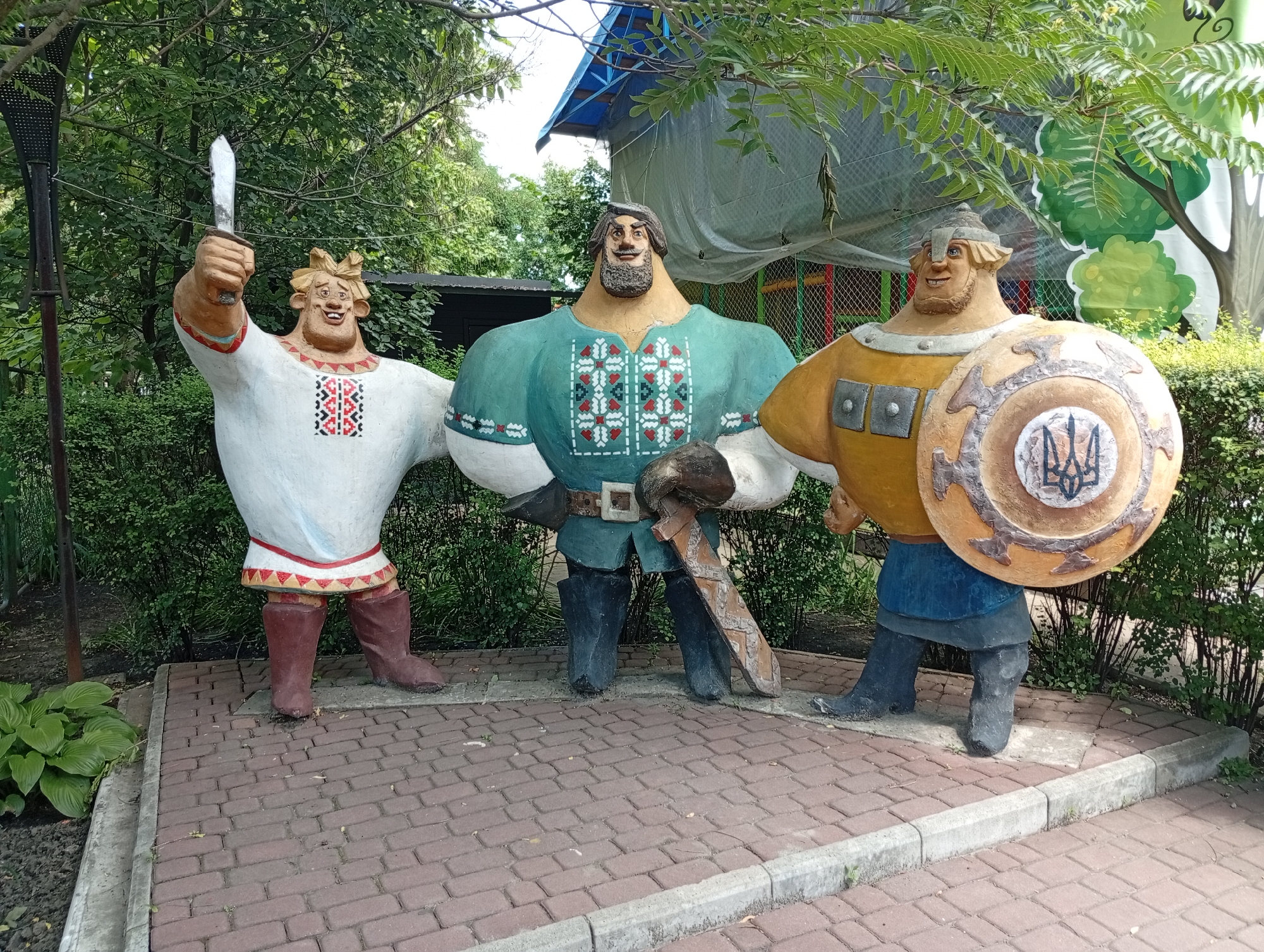 Three large, cartoonish statues of men stand in an outdoor setting on a paved walkway. The statue on the left is dressed in a white tunic with red and black embroidered trim around the neckline and hem, and brown boots. He holds a silver knife aloft in his right hand. The central statue wears a teal tunic with a similar embroidered pattern around the neckline, a brown belt, and black boots. He holds a large, brown sword horizontally in front of him. The statue on the right is clad in a yellow tunic, blue pants, and dark boots. He carries a large, round shield on his left arm, which features a blue and black trident symbol. All three statues have exaggerated features, with broad shoulders, large heads, and jovial expressions. They are positioned in front of lush green bushes and trees, with a blurred building and a blue tarp visible in the background. The ground is covered in reddish-brown brick pavers with a concrete curb bordering the walkway.
