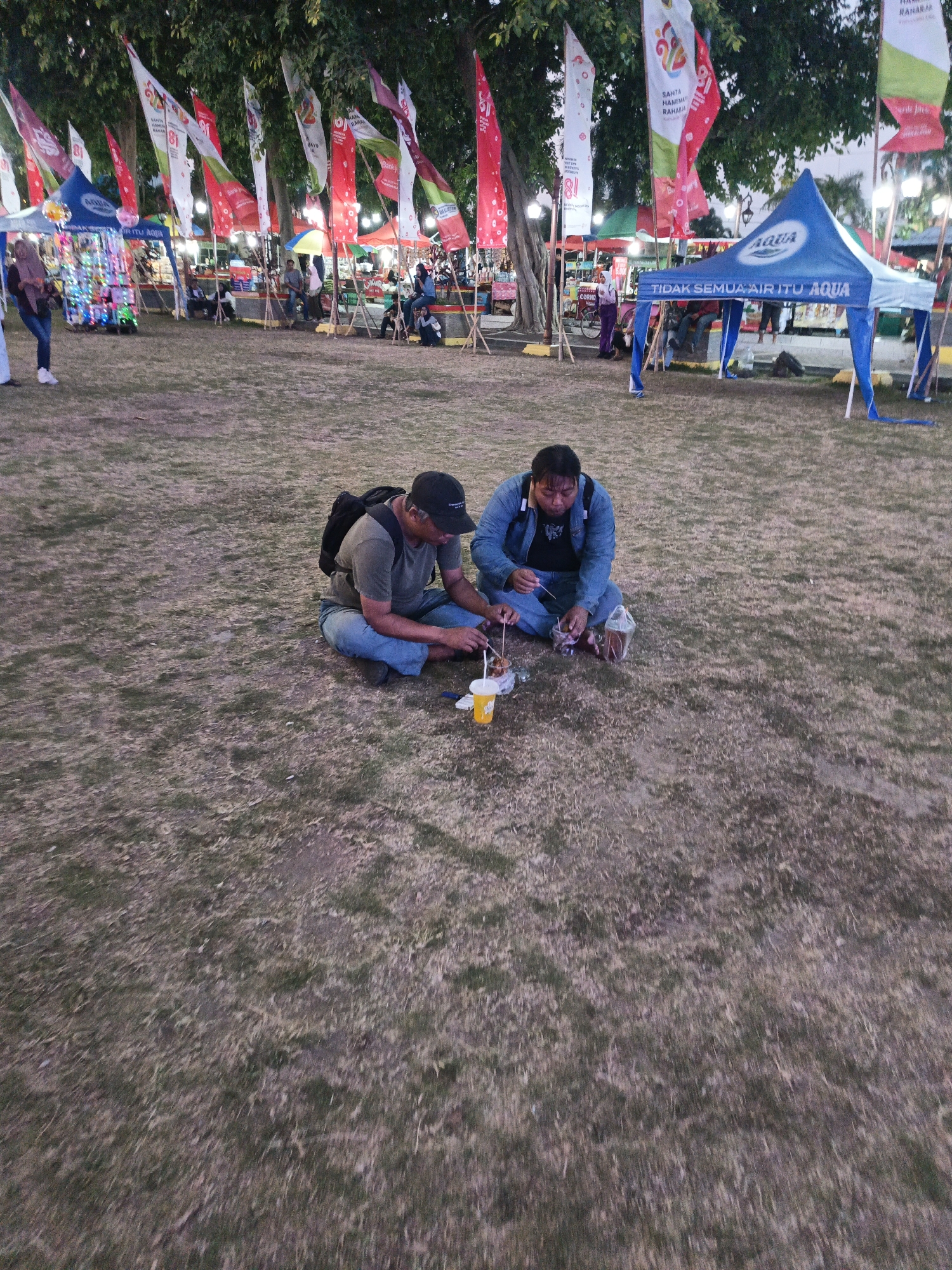 Two men are sitting on the ground in a park area at dusk in Klaten, Indonesia. They are both facing each other and appear to be eating. One man is wearing a black baseball cap, a dark t-shirt, and denim shorts. He has a backpack on. The other man is wearing a blue denim jacket over a dark t-shirt and blue jeans. They are both holding skewers and there are some food items and drinks on the ground near them. In the background, there are several tents and flags, suggesting a market or festival. The ground is covered in dry grass. The overall lighting indicates it is late afternoon or early evening.