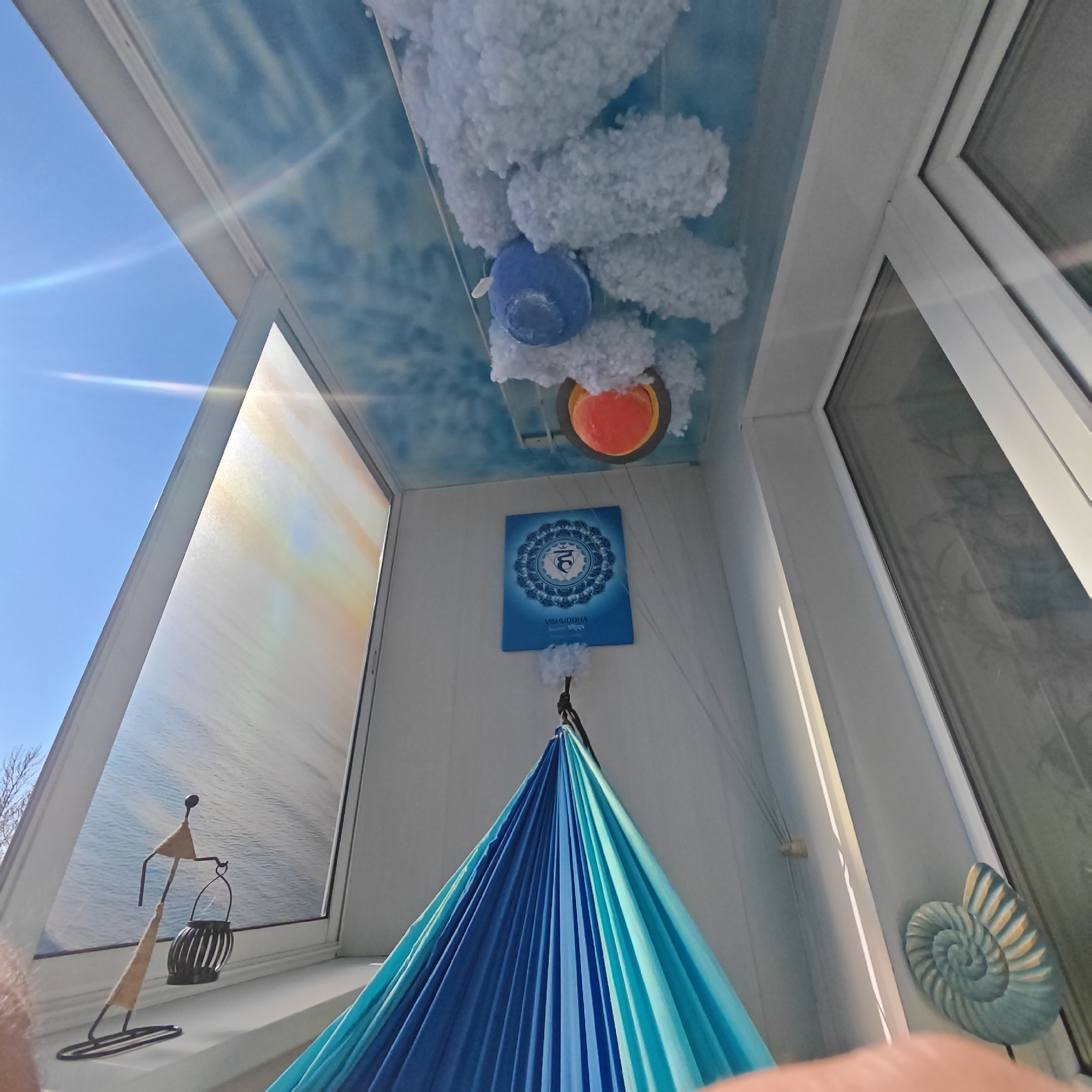 The image shows an interior space, possibly a balcony or enclosed porch, with a whimsical and creative design. The ceiling is painted with a blue sky and cloud motif and has three-dimensional cloud-like decorations hanging from it, along with a blue and an orange decorative object. A blue banner featuring the Vishuddha chakra symbol is hung on the wall. A blue and turquoise hammock is suspended from the ceiling. A metal sculpture depicting a figure with a lantern sits on a windowsill, next to a stylized image of waves. A nautilus shell decoration is placed on the adjacent wall. The windows show a view of a clear blue sky. No people are visible in the image. The location in the city of Rostov, Russia, cannot be determined from the visual content.