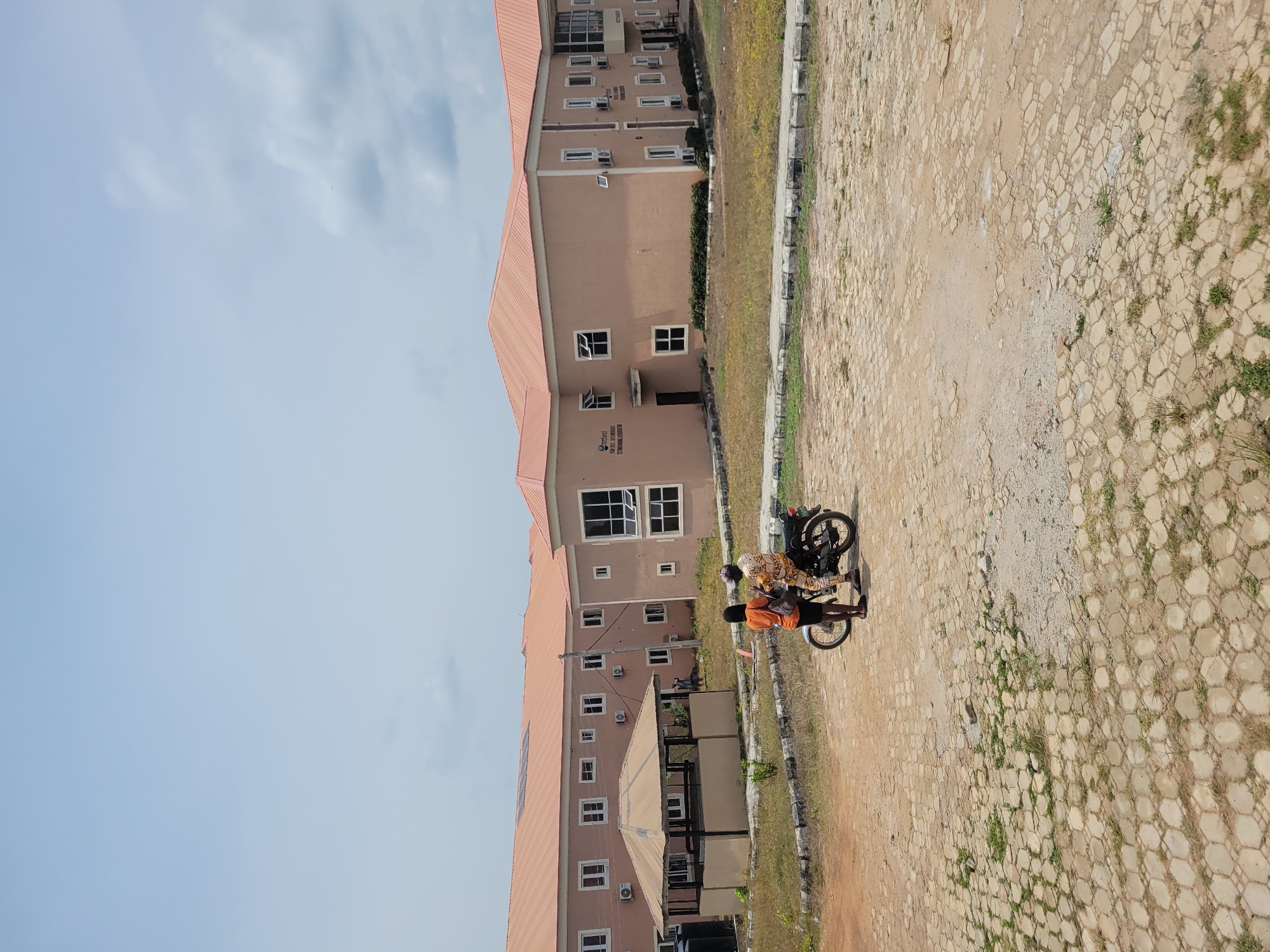 Two people are standing next to a motorcycle in a paved area in front of a modern building complex. The building has a tan facade and a reddish-orange roof. There are multiple windows and air conditioning units visible on the building. To the left of the building, there is a covered outdoor seating area with a tan roof. The ground is a mix of pavement and dirt. The sky is overcast.