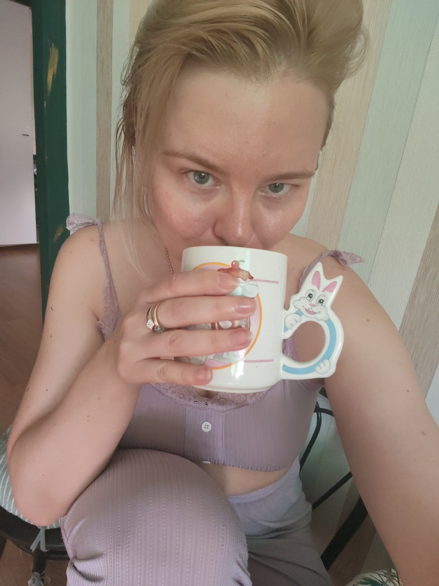 A fair-skinned woman with blonde hair is drinking from a mug. The mug is white with a cartoon image of a sundae and a rabbit-shaped handle. The woman is wearing a light purple top with lace trim. She has two rings on her left hand. She is sitting on a chair with a striped seat. The background is a wall with vertical stripes of varying colors. A dark green door frame is visible in the left corner. The image does not provide sufficient information to confirm the location is Izhevsk, Russia.