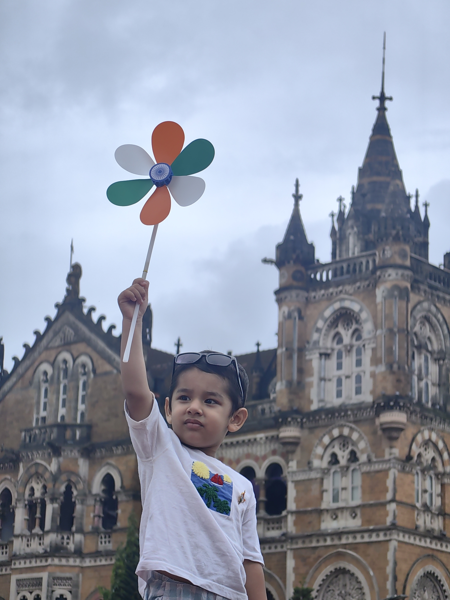 A young boy, likely of Indian ethnicity, stands outdoors holding a pinwheel aloft in his right hand against a cloudy sky. The pinwheel has petals colored orange, green, and white, with a blue central disk featuring a depiction of the Ashoka Chakra, representing the Indian flag. The boy is wearing a white t-shirt with a colorful graphic and sunglasses perched on his head. In the background, a large, ornate building with gothic architectural elements, characterized by multiple spires, arched windows, and intricate stonework, is visible. This building is identified as Chhatrapati Shivaji Maharaj Terminus (formerly Victoria Terminus), a historic railway station and UNESCO World Heritage Site located in Mumbai, India. The overall scene suggests a patriotic or celebratory event, possibly Independence Day in India, given the colors and symbolism of the pinwheel.