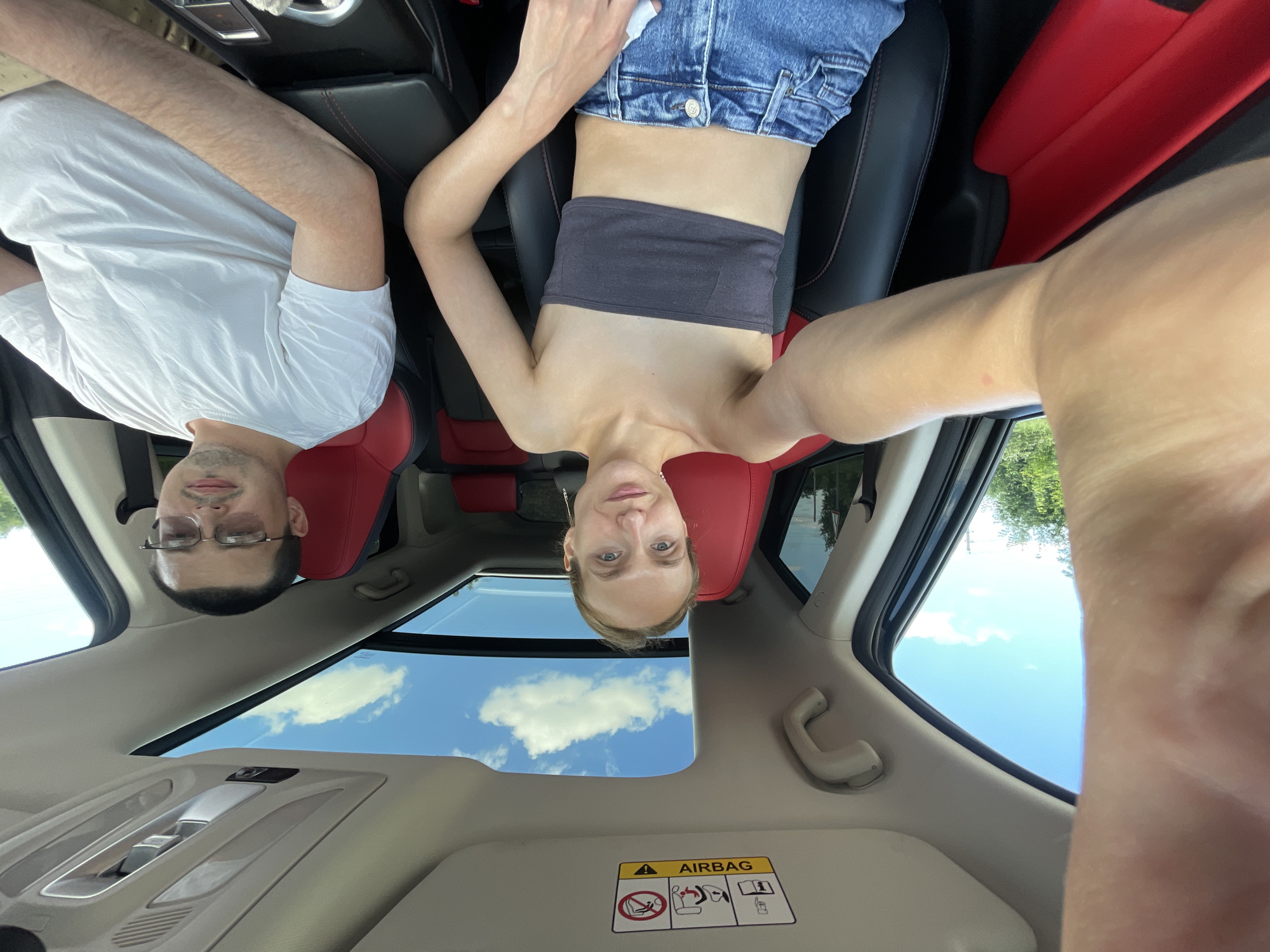 A front-facing, eye-level selfie is captured from the perspective of the passenger seat of a car. A young woman with blonde hair pulled back and no makeup is in the passenger seat, wearing a dark gray bandeau top and blue jeans. To her right, in the driver's seat, a man wearing glasses and a white t-shirt is visible. The car's interior features red and black leather seats and a panoramic sunroof, with a blue sky and clouds visible through it. The image appears to have been taken during the daytime, with ample natural light. The car is possibly located in the city of Kuyeda, Russia, given the context provided, though this cannot be definitively confirmed from the image alone.