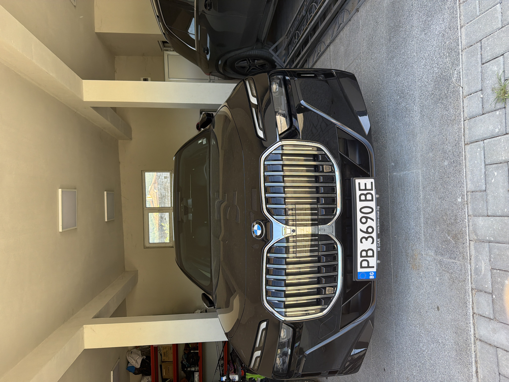 A black BMW car is parked in what appears to be a garage or carport. Another dark-colored vehicle is partially visible behind and above it. The car has a Bulgarian license plate, "PB 3690 BE," which would indicate the license plate is registered in the city of Plovdiv, Bulgaria. Based solely on the location of the license plate and lacking any specific environmental clues within the image, it is impossible to determine if the location is within the city of Asenovgrad or elsewhere. The garage/carport is painted in light tones, and its internal space includes storage areas visible in the background, indicating a space for storing items as well as parking vehicles. The floor next to the car appears to be paved with grey stone tiles or blocks. The area appears to be enclosed, shielded from external weather conditions.