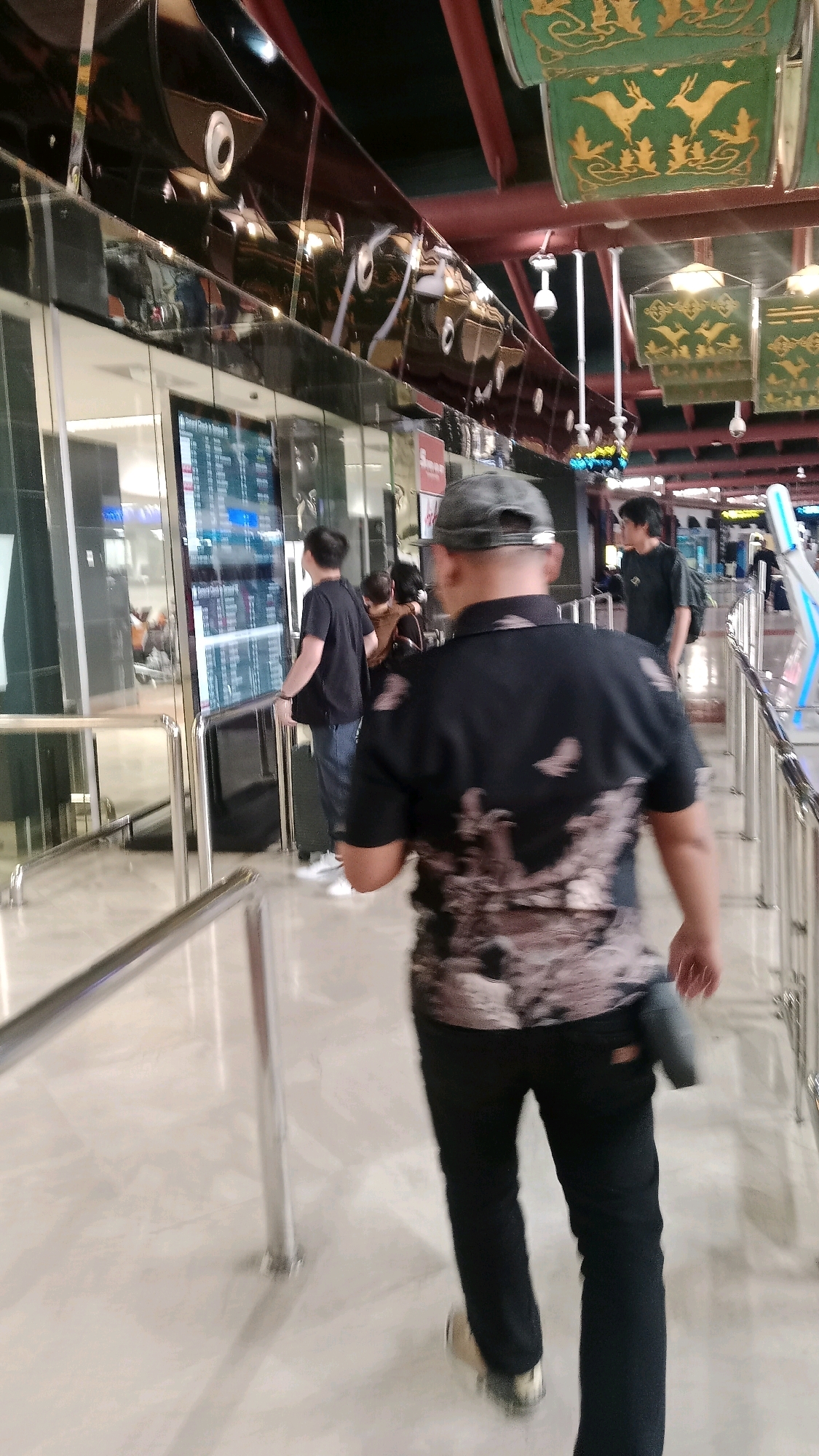 The image shows the interior of an airport terminal. In the foreground, a person wearing a black patterned shirt and black pants walks away from the camera. The person has a grey cap on and a dark object attached to their right hip. In the background, several other people are visible, some looking at a large digital display screen showing flight information. There are metal railings, a polished floor, and decorative elements hanging from the ceiling, one of which features images of deer. The environment suggests a public transportation hub, likely an airport, and the context indicates it is located in Tangerang, Indonesia.