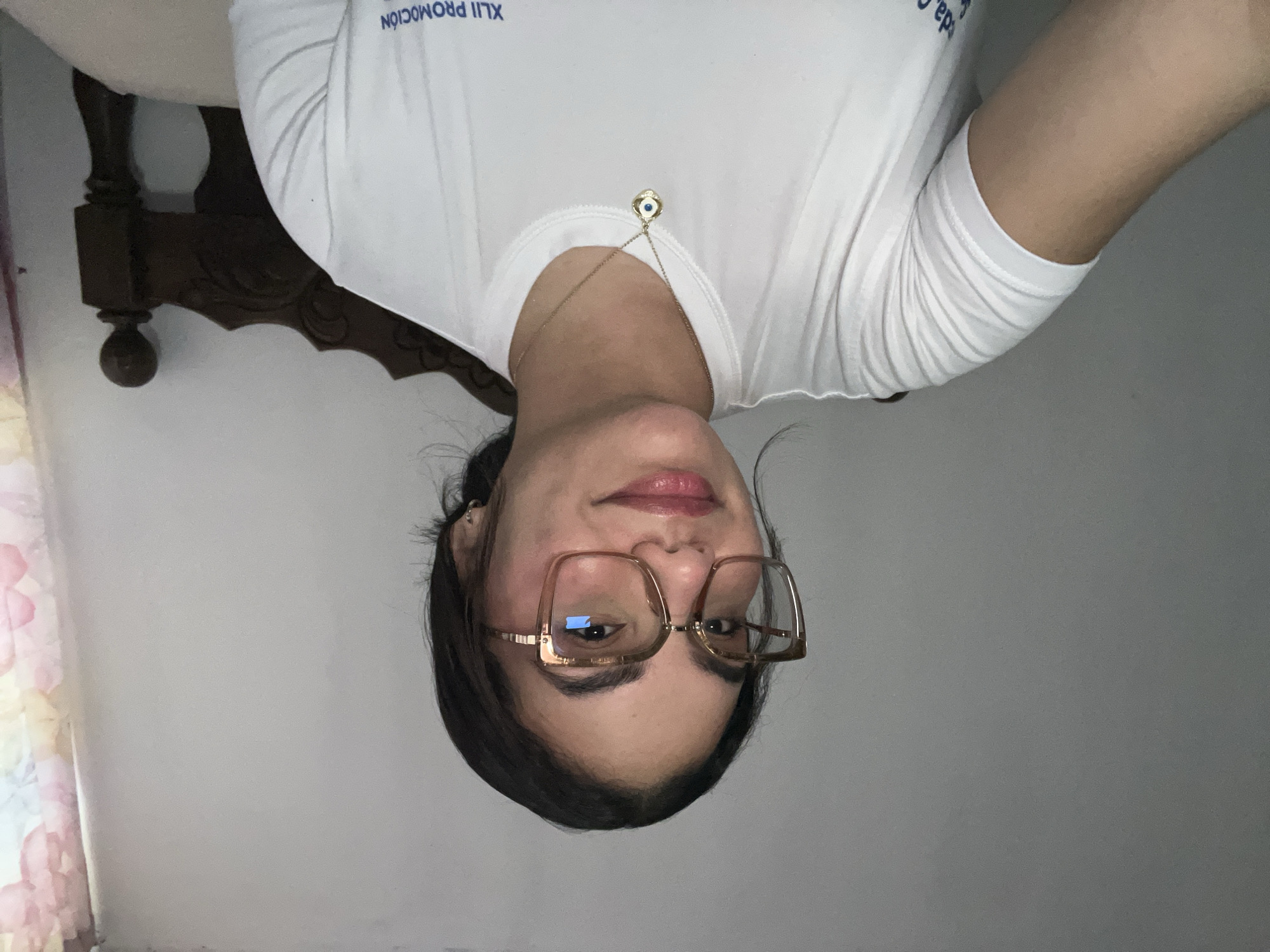 A young woman with dark hair and glasses is looking at the camera, wearing a white t-shirt with a gold necklace. The background is a plain wall with some floral curtains to the right. She appears to be in a bedroom. The text on her shirt is partially visible, with "XLII PROMOCION" and some other Spanish words.