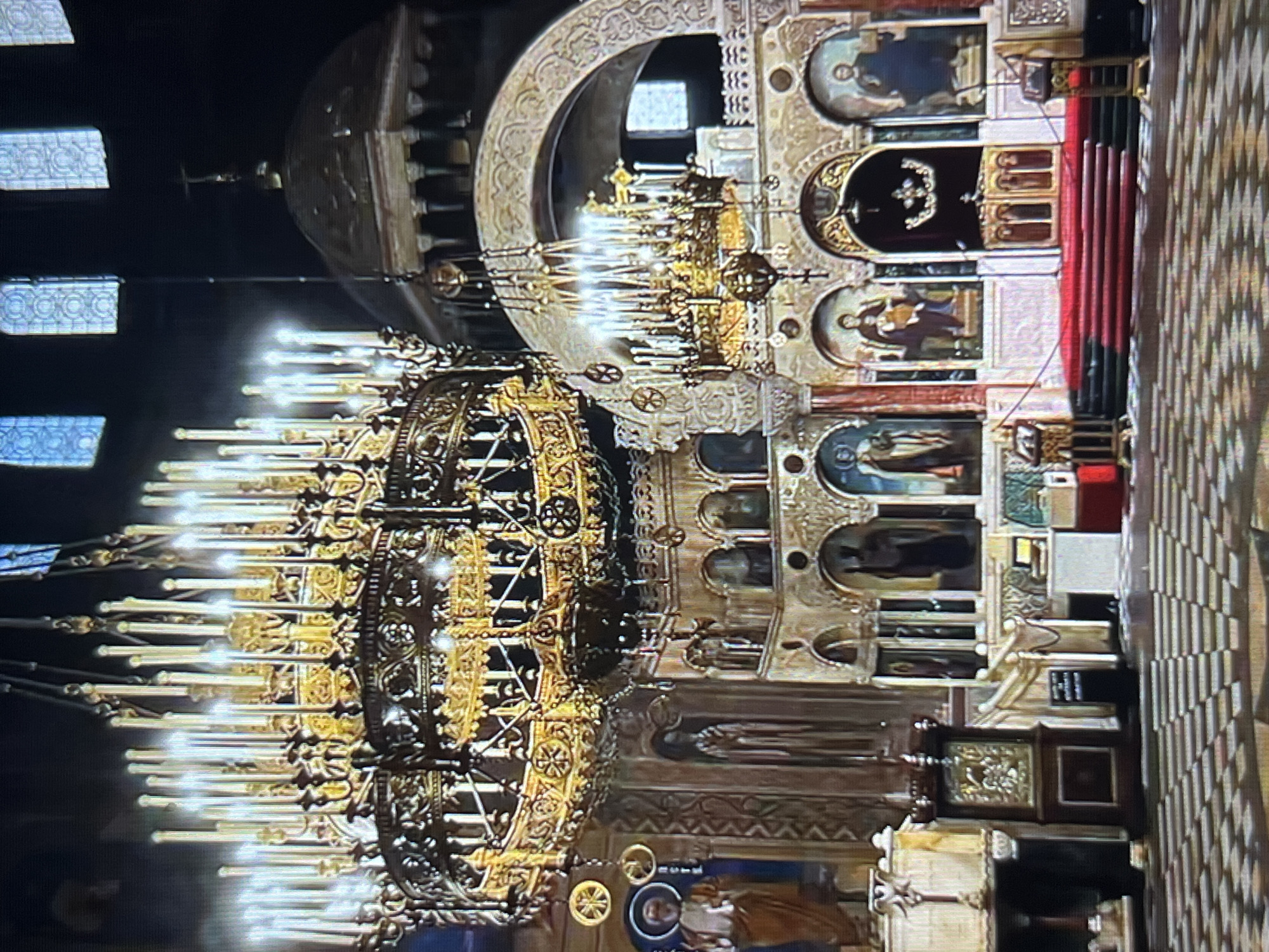 The image captures the interior of a church, likely an Orthodox or Eastern Catholic church, given the ornate iconography, chandeliers, and architectural style. The chandeliers are large and complex, featuring numerous lights resembling candles. The walls are decorated with religious paintings in arched recesses. A red carpet covers steps leading up to an altar or sanctuary area. The flooring has a checkered pattern. The interior has a dark, dimly lit atmosphere, illuminated primarily by the chandeliers. The church is of unknown location in Tinaquillo, Venezuela. No people are visible.