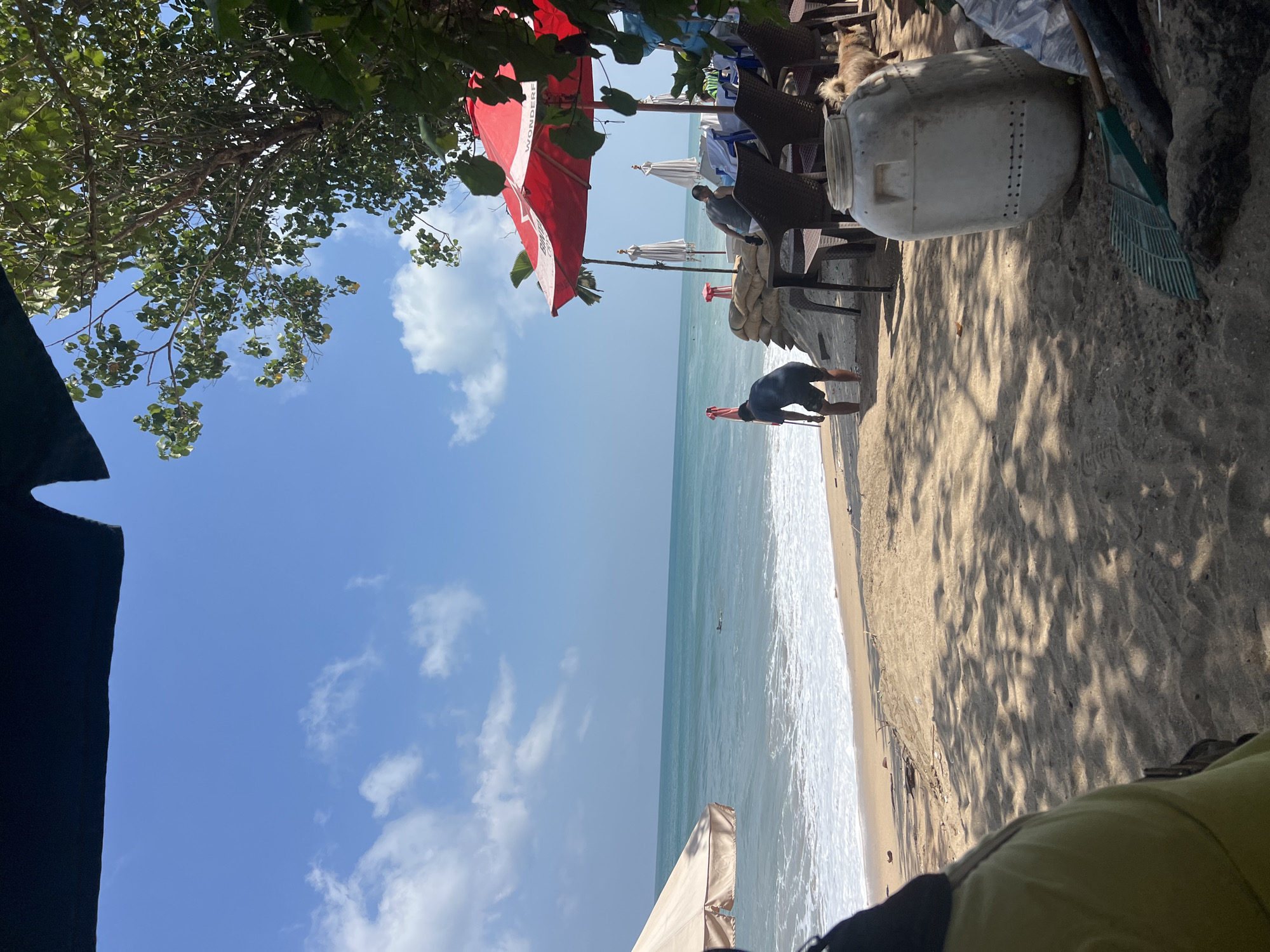 A tranquil scene unfolds on a sun-drenched beach in Kuta, Indonesia. The turquoise ocean stretches to the horizon, meeting a sky adorned with fluffy white clouds. Gentle waves crash onto the sandy shore, leaving a frothy white surf. In the foreground, a person in dark shorts and a blue shirt is bent over, seemingly tending to something near the water's edge. Further back, under the shade of a large red umbrella, two individuals are seated, while another person stands nearby. Sandbags are piled up, forming a protective barrier. Several white beach umbrellas stand at attention, hinting at a popular gathering spot. A large white plastic container, perforated with holes, sits on the sand, accompanied by a rake, suggesting some form of cleanup or maintenance. A small, fluffy dog can be seen in the background, adding a touch of domesticity to the natural setting. The scene is framed by the dappled sunlight filtering through the leaves of a lush tree.