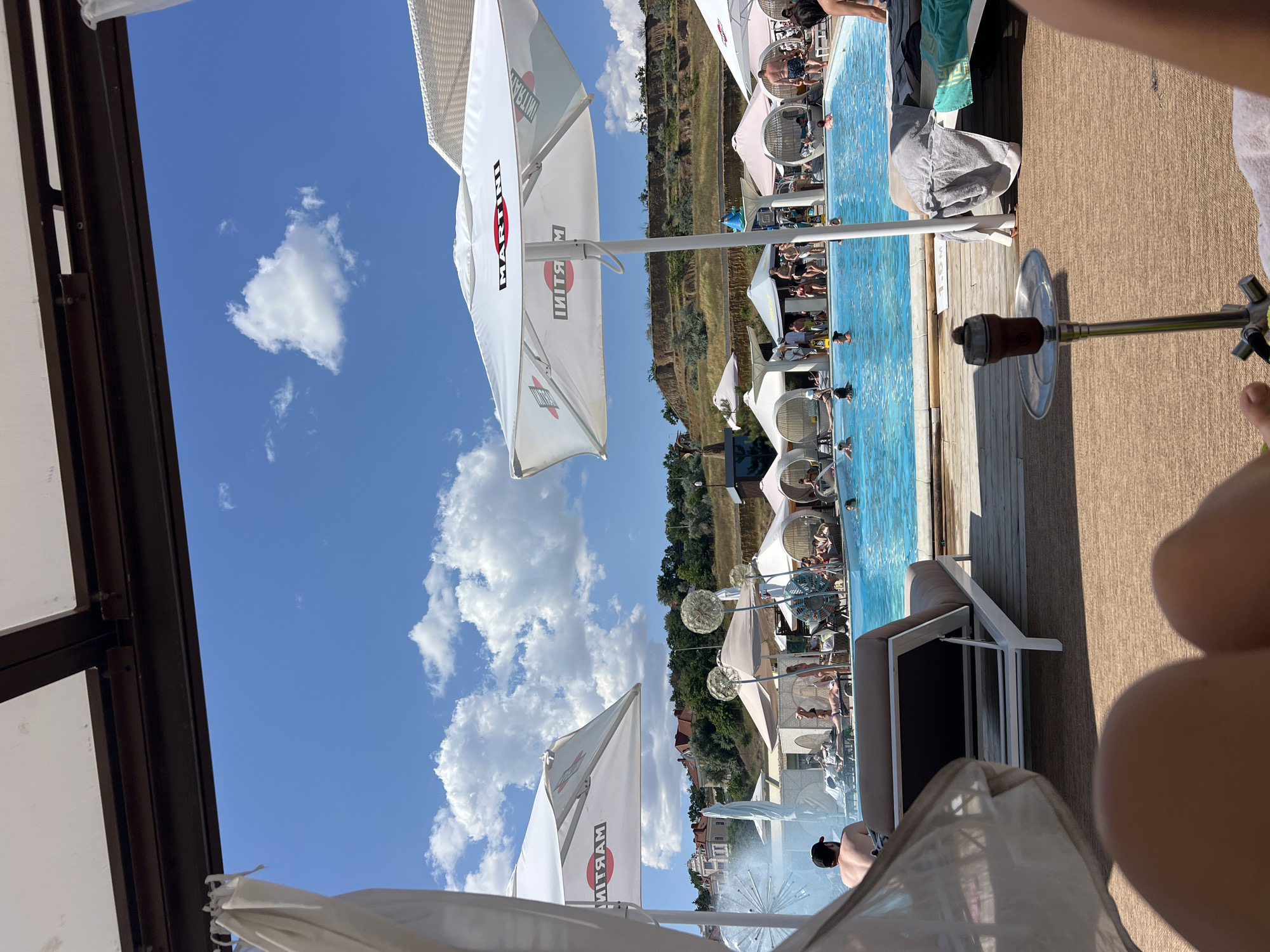 This is a ground-level, eye-level, long shot of a bustling outdoor swimming pool area with many people enjoying the water and sun. The scene is captured on a sunny day with a bright blue sky dotted with fluffy white clouds. In the foreground, a hookah pipe is prominently placed on a beige, woven mat, with a person's legs and feet visible to the right, suggesting they are relaxing.
A white lounge chair with a brown cushion is positioned to the left, with a person wearing a black baseball cap facing away from the camera, looking towards the pool. The pool itself is a vibrant blue, with several people swimming and lounging in and around it. Various white cabanas and umbrellas, some with the "MARTINI" logo visible, are scattered around the pool area, providing shade.
In the background, a lush green hillside is visible, with buildings and trees dotting its slopes. The architecture of the buildings suggests a resort or a developed coastal area. A water fountain with a spray of water is also visible to the left of the pool. The overall atmosphere appears to be one of leisure and recreation, typical of a beach club or resort. The location is identified as Fontanka, Ukraine.