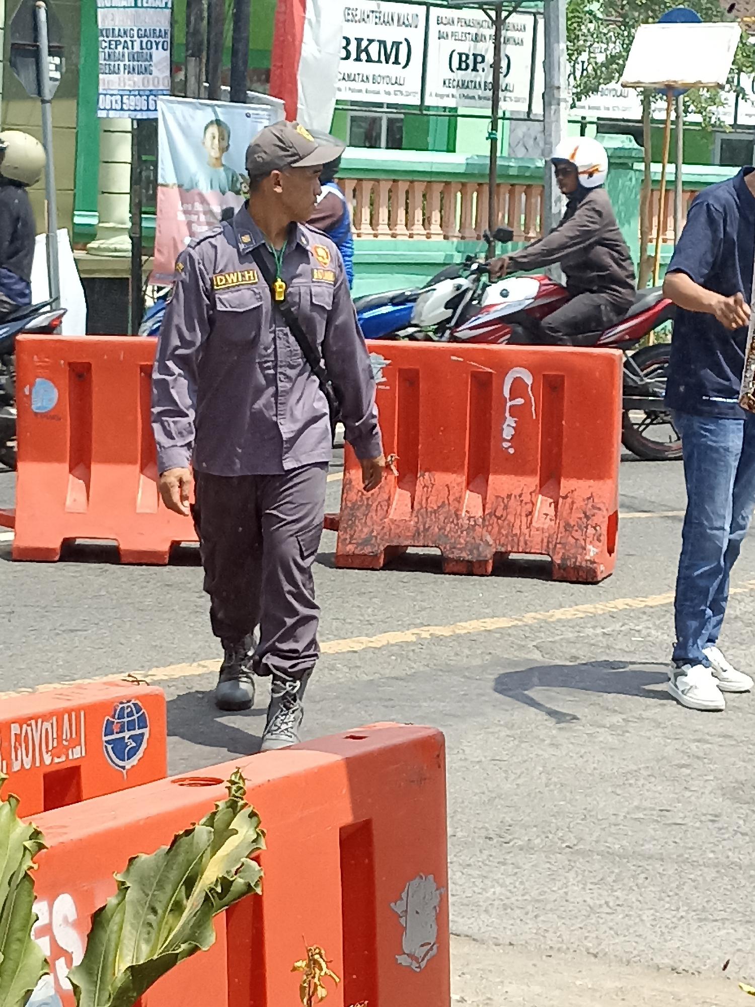The image depicts an outdoor scene with several people and objects on a street in Pulisen, Indonesia. In the foreground, a man wearing a dark gray uniform, a cap, and dark boots walks towards the viewer's left. He has a name tag on his uniform that reads "IDWH". He is carrying a dark bag slung across his chest. To his right, a section of orange plastic traffic barrier is visible. Behind the barrier, a motorcycle with a rider wearing a white helmet and a dark jacket is seen. Further in the background, another traffic barrier and a person in a dark blue shirt and jeans are visible. A street sign with text in Indonesian is present on a pole in the background. The ground appears to be asphalt, with yellow road markings visible. The environment suggests a daytime scene, with sunlight casting shadows.