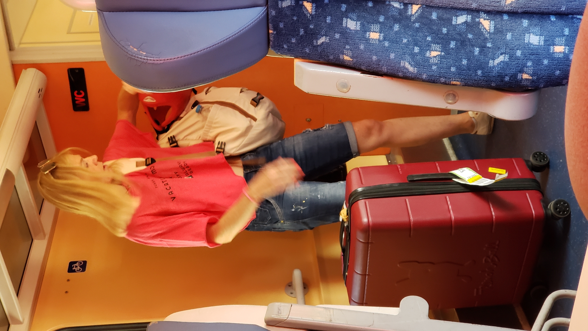 A blonde woman wearing sunglasses on her head and a pink t-shirt stands on a train. She has a tan bag on her shoulder and is reaching into it. She is wearing denim shorts and white sneakers. In front of her is a red suitcase. The train car is orange and blue. There is a WC sign on the wall and a bicycle symbol. There are seats in the foreground.