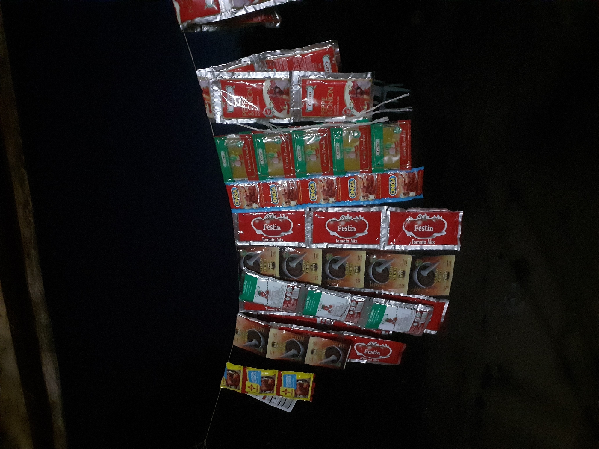 In Jalingo, Nigeria, a string is strung between two points, holding numerous small packets of spices. The packets are varied in color, with some being red, yellow, green, and silver. Some packets are stacked in columns, while others hang individually. The background is dark, suggesting it is nighttime or a dimly lit area. The packets appear to be of different brands and contain various spice mixes, including curry powder, pepper, and onion seasoning mix. The scene is outdoors, possibly at a market stall or a roadside vendor, with a dark, unidentifiable surface in the foreground.