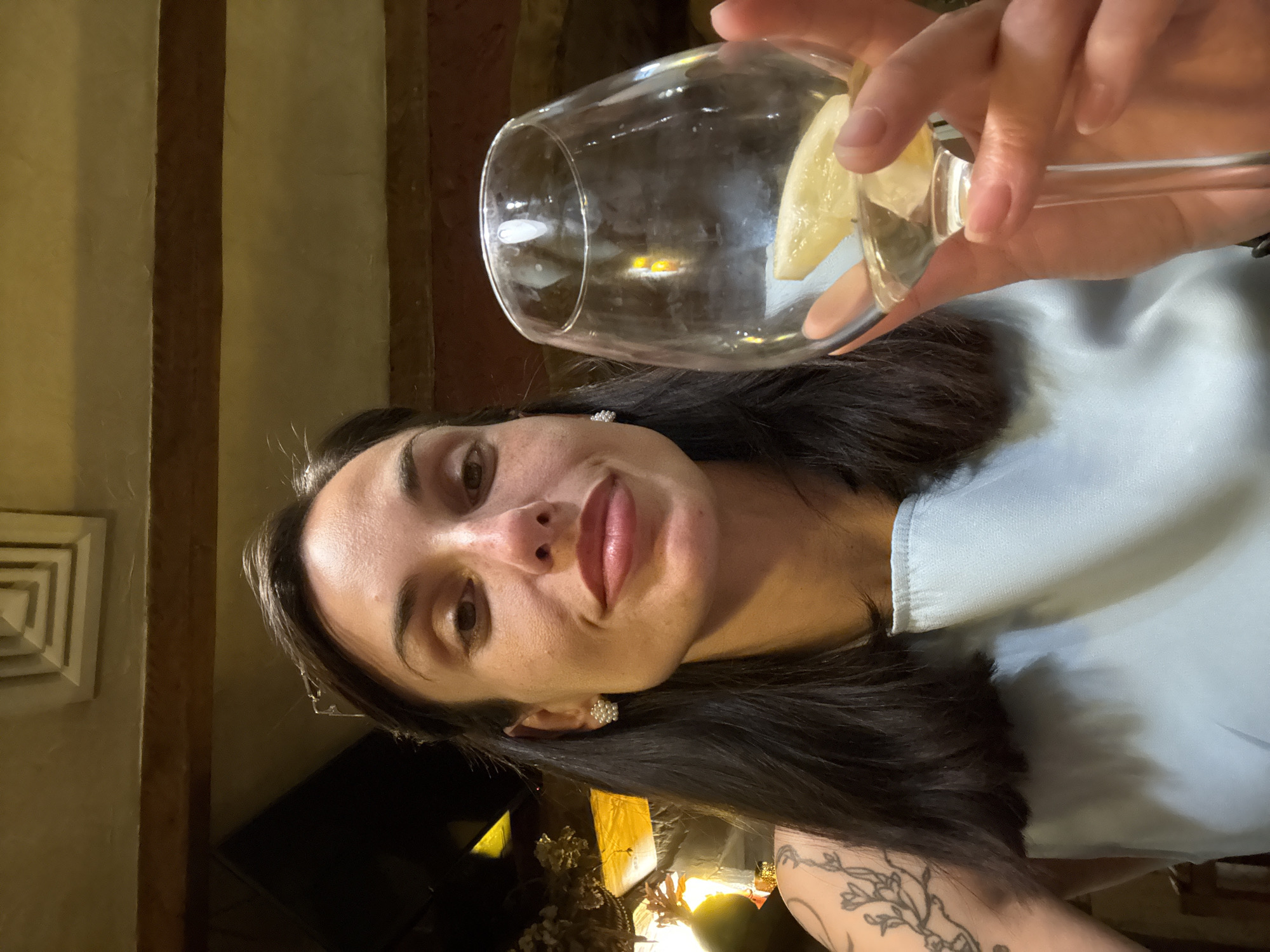 A woman holds a wine glass containing a lemon wedge. She has dark hair and is wearing a light blue top. The background appears to be a dimly lit indoor setting, possibly a restaurant or bar. Based on the prompt, the location is in the city of Pleven, Bulgaria.